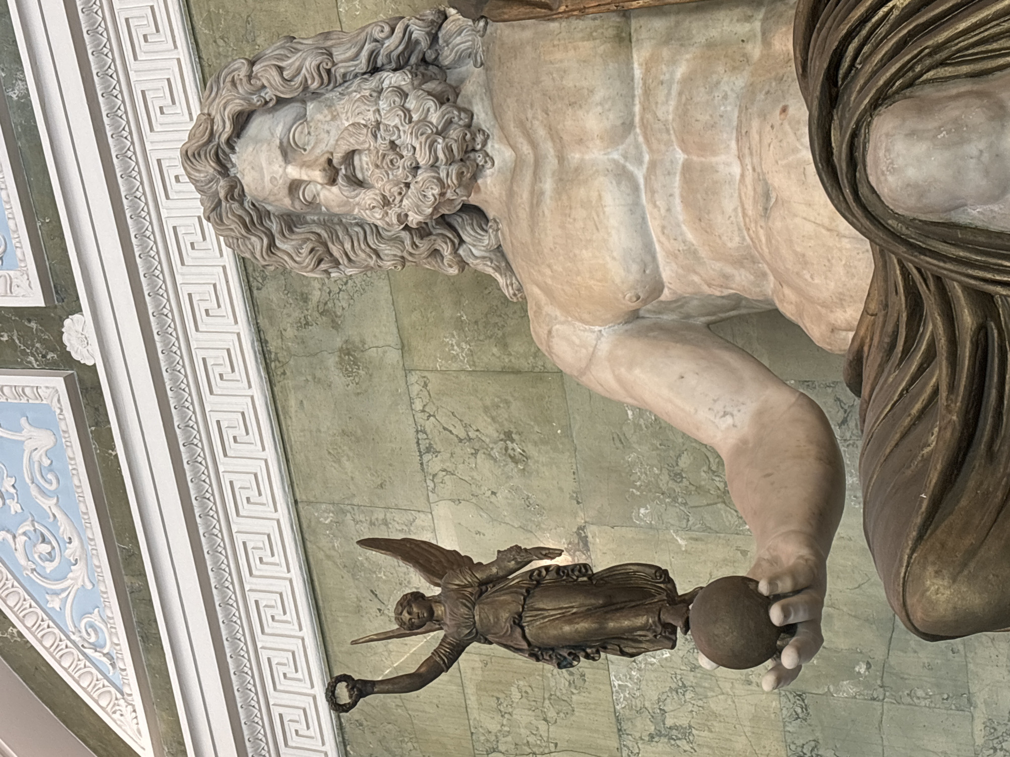 This media file shows a close-up, low-angle view of a monumental marble sculpture, likely depicting a Greek or Roman deity, possibly Jupiter or Zeus, given the muscular physique and flowing beard. The sculpture is predominantly made of marble, with a draped garment of a darker, bronze-like material. The figure's head, with its detailed curly hair and beard, is tilted upwards and to the left. The torso is powerful and chiseled, showcasing anatomical detail.
In the foreground, held by the sculpture's right hand, is a small, dark bronze statuette of a winged figure, possibly Nike, the goddess of victory. This winged figure is holding a wreath in its left hand and what appears to be an orb in its right. The background consists of a green, marble-patterned wall and ornate white architectural moldings, featuring Greek key patterns and decorative relief work. The lighting suggests an indoor setting, possibly a museum or grand hall, with diffused light illuminating the sculptures. The image has been rotated 90 degrees clockwise, so the subjects are oriented sideways.
The scene appears to be a display of classical art, possibly within a museum or a grand architectural space in Saint Petersburg, Russia. There are no visible signs of weather or time of day, as the setting is indoors. The overall emotion conveyed is one of grandeur and artistic admiration. No text is visible in the image.
