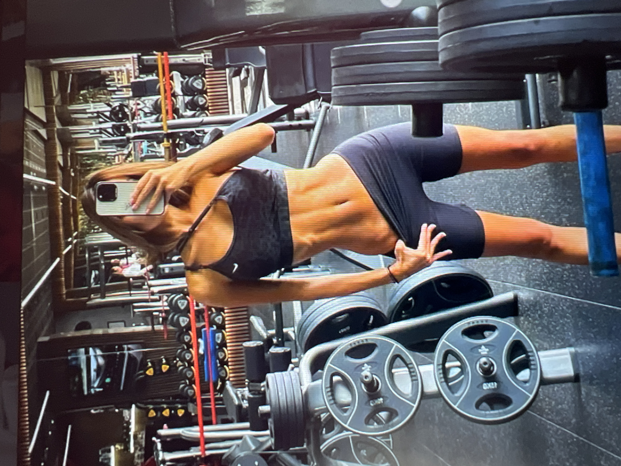 The image shows a woman taking a mirror selfie in a gym. She is wearing a sports bra and shorts, and her midriff is exposed, revealing abdominal muscles. The gym appears to be well-equipped with weight machines, dumbbells, and weight plates. The environment is dimly lit with artificial lighting. The background, seen in the mirror, shows rows of gym equipment and what appears to be other people in the distance, also exercising. The gym's aesthetic suggests a modern and professional setting. The location is identified as the city of Sofia, Bulgaria.