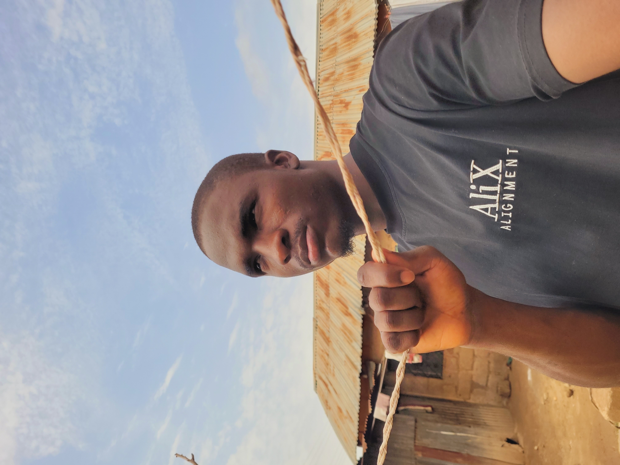 A man in a black shirt with the words "Alix Alignment" on it is holding a rope in his right hand, with a corrugated metal roof of a building visible behind him, and the sky above. The photograph was taken outdoors in the city of Bukuru, Nigeria.