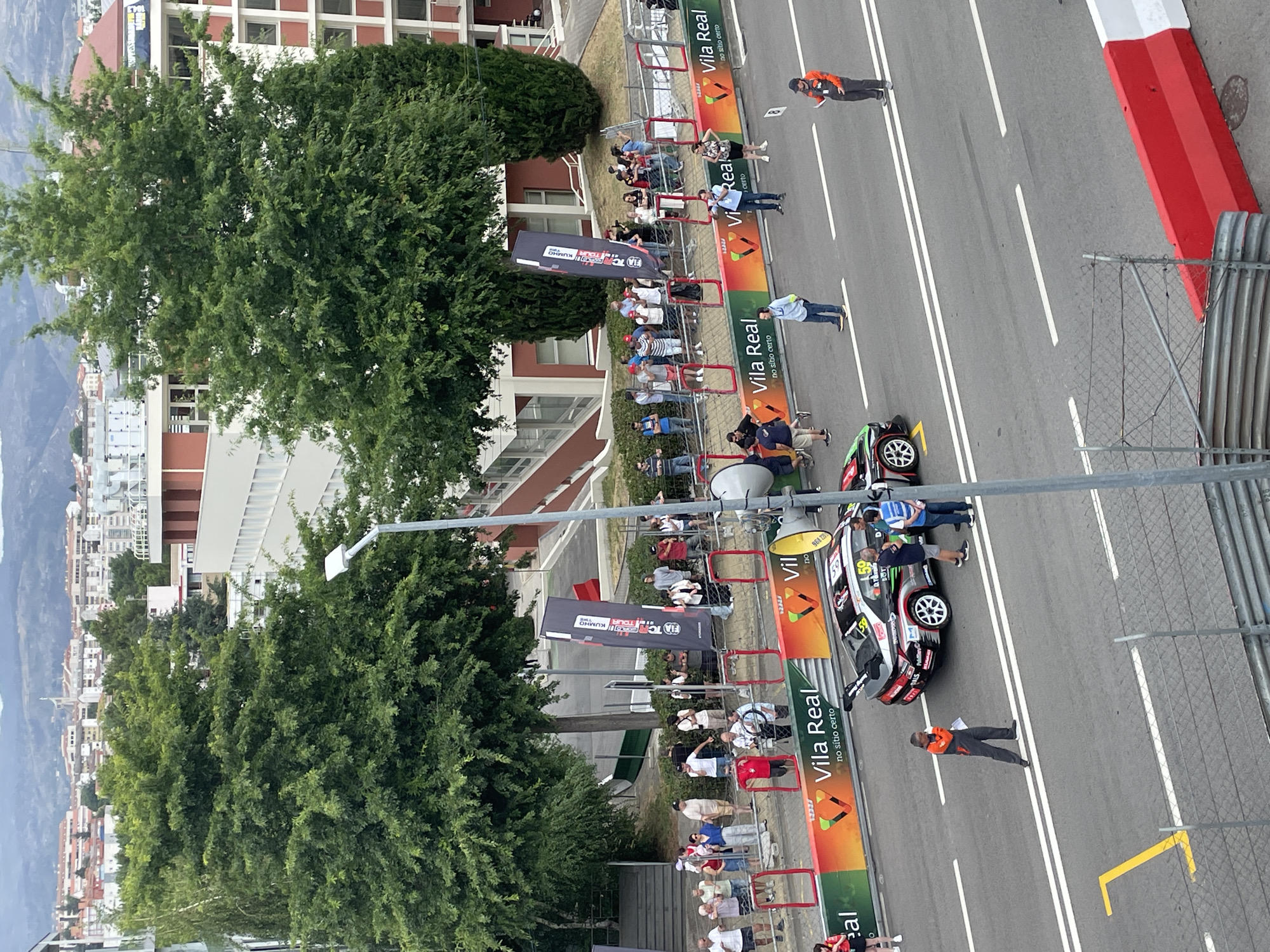 A high-angle, slightly overcast outdoor shot captures a street circuit race event in Vila Real, Portugal. In the foreground, a race car with the number 59 is parked on the asphalt, its rear wing prominently displayed. Several individuals are gathered around the car, including a person in an orange and grey racing suit holding a clipboard, seemingly supervising. Further up the track, another person in a similar suit stands observing. Spectators line the barriers along the roadside, some holding phones to take pictures, while others converse. Large banners with the "Vila Real" logo and "no sítio certo" text are displayed along the track. In the background, lush green trees rise above the barriers, and beyond them, a cityscape with various buildings and distant hills is visible. A tall street lamp stands near the trees. The overall scene depicts a paused moment during a racing event, with participants and onlookers present.