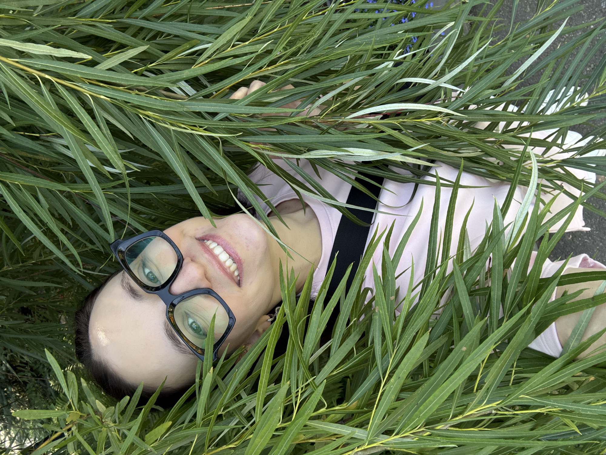 A young woman with dark hair and glasses is lying down amidst dense green foliage, her head tilted up towards the camera. She is smiling, with her teeth showing, and her eyes are visible behind her large, dark-rimmed glasses. She is wearing a light pink t-shirt with a black strap visible across it. The foliage is lush and green, with long, thin leaves creating a natural frame around her face. The background is obscured by the leaves. The lighting suggests an outdoor setting, likely during the day. The image appears to be a selfie. There is no information that can ascertain the location as Chaikovsky, Russia.