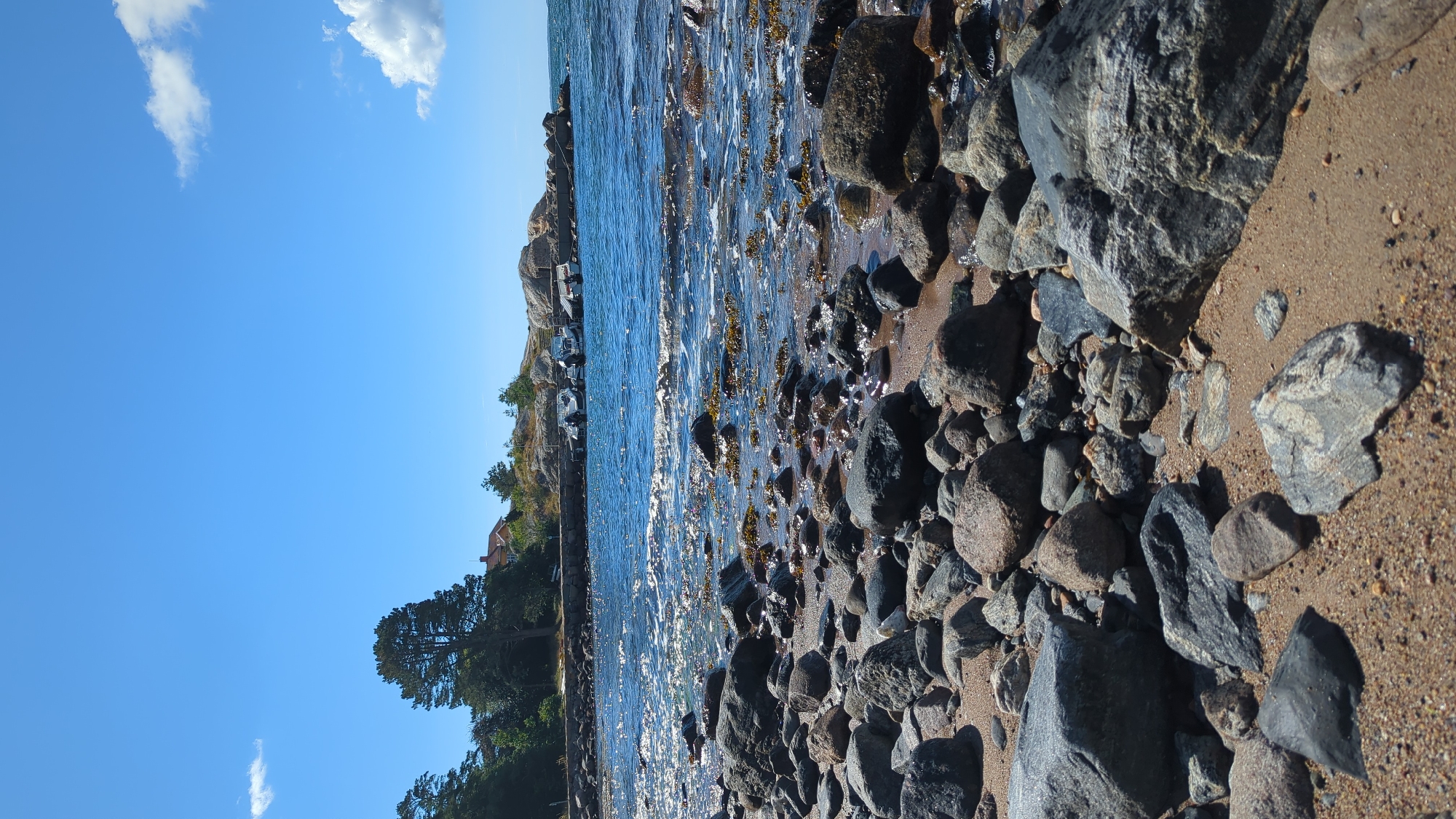 This image depicts a rocky shoreline with calm blue water under a clear sky with scattered white clouds. In the foreground, numerous grey and brown rocks of various sizes are scattered on a sandy beach. The water gently laps at the shore, with some seaweed visible. In the background, a line of trees and a house with a reddish-brown roof are visible on a slight incline. Several motorboats are docked along a pier or rocky outcropping in the middle ground. The scene is set in Larkollen, Norway.
