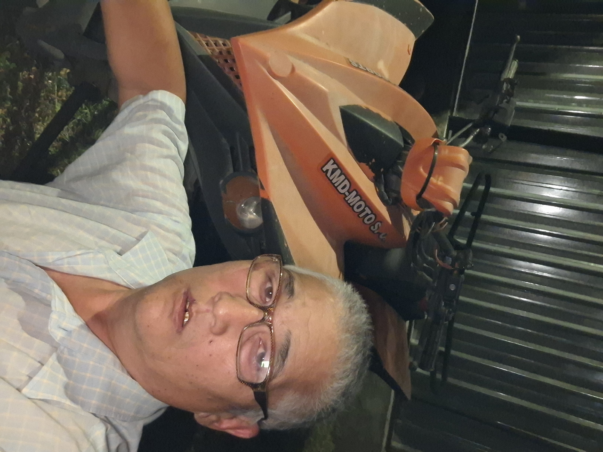 The media file shows a close-up of a man with graying hair and glasses, wearing a light blue checkered shirt. He is looking towards the camera with his mouth slightly open, revealing some gold teeth. To his right, an orange and black motorcycle is visible. The motorcycle has the text "KMD-MOTO S.C." printed on its side. The scene appears to be outdoors, possibly in a residential area or a parking lot, with a corrugated metal fence in the background. The lighting suggests it might be evening or nighttime. The man's expression does not convey any strong emotion. There are no clear weather cues. Given the location context of Almaty, Kazakhstan, the scene could be any urban or semi-urban setting within the city.
