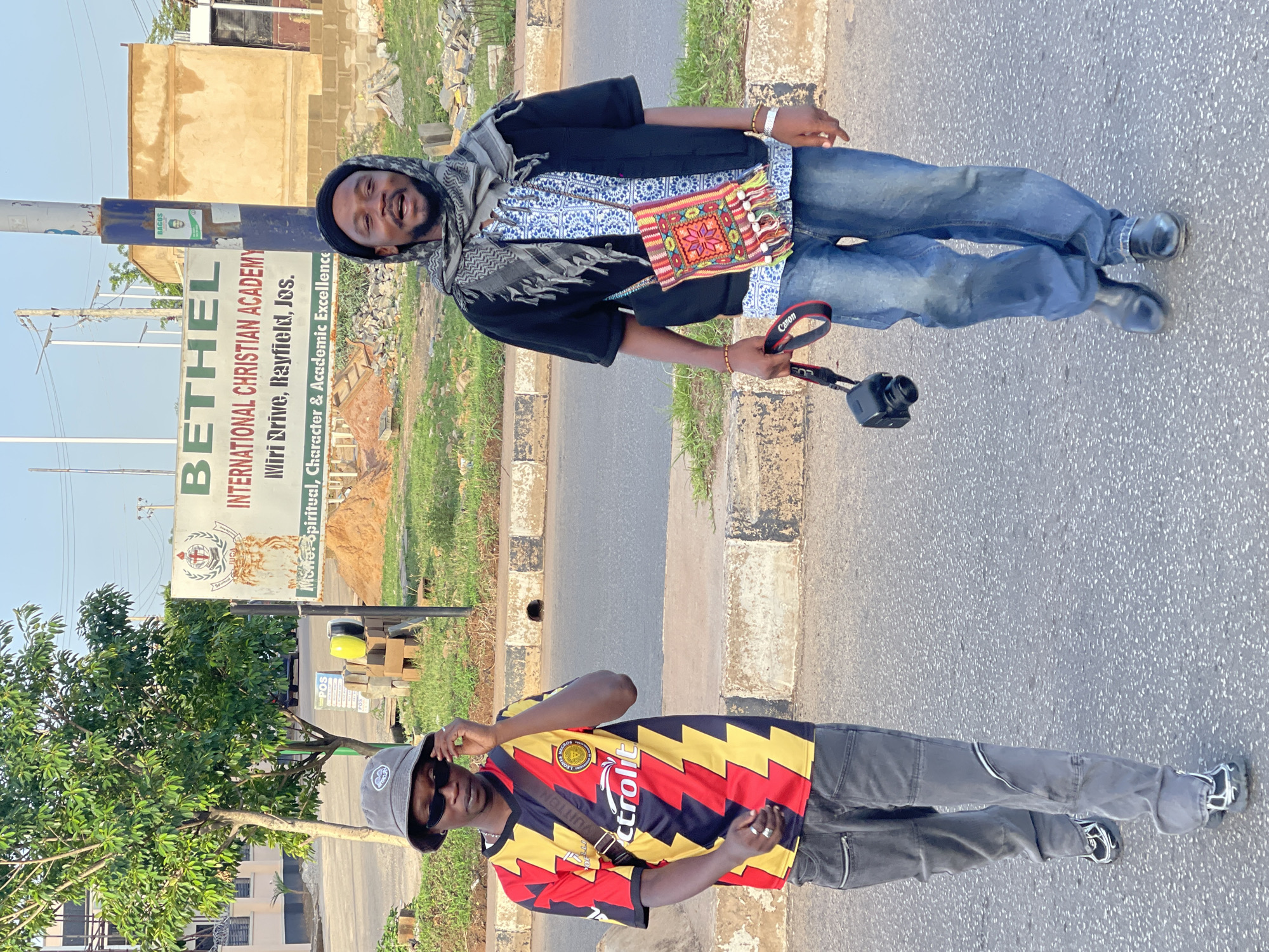 Two young men are standing beside a street in what appears to be Bar Jirgi Summa, Nigeria. One is wearing a grey and black patterned head covering, a scarf, a black coat over a patterned shirt, blue jeans, and black shoes. He carries a colorful bag and a Canon camera. The second man, positioned lower in the image, wears sunglasses, a grey bucket hat, and a soccer jersey with black, red, and yellow lightning bolt patterns. He is also wearing grey pants and light-colored athletic shoes. In the background is a sign for "Bethel International Christian Academy, Miri Drive, Rayfield, Jos." The environment appears to be a residential street with buildings and vegetation in the backdrop. The image is horizontally oriented.
