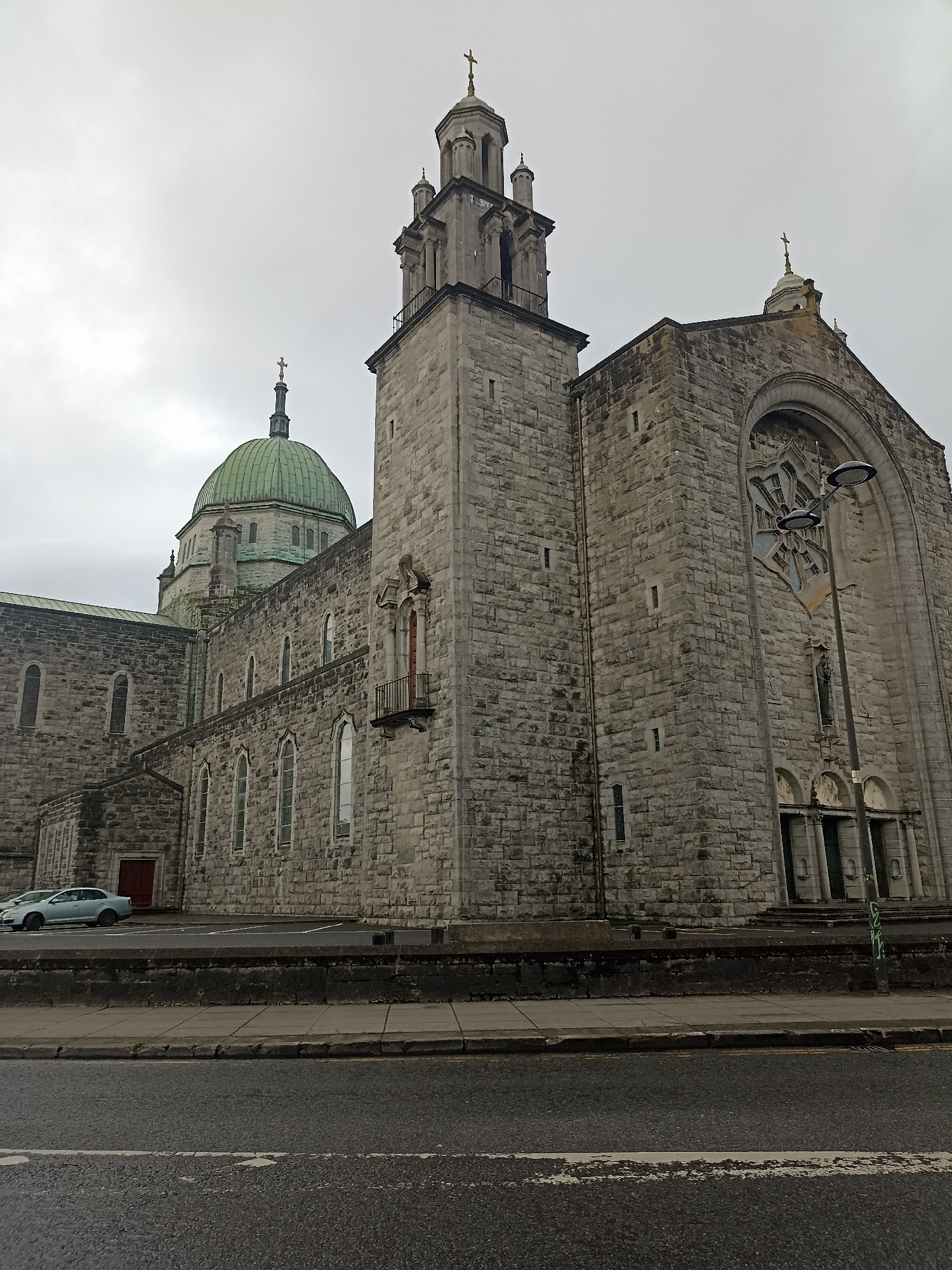The image shows a view of the Galway Cathedral, a Roman Catholic cathedral located in the city of Galway, Ireland. The cathedral is a large stone building with a prominent green dome and a tall bell tower. The facade features arched windows and a large rose window above the main entrance. A street with parked cars and a sidewalk runs in front of the cathedral. The sky is overcast, suggesting a cloudy or rainy day.
