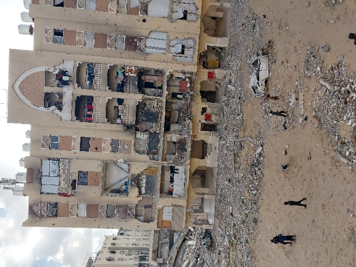 The image depicts a scene in Khan Yunis, Palestinian Territory. A severely damaged multi-story building with a tan-colored facade dominates the central view. This structure exhibits extensive damage, including numerous circular impact marks consistent with bullet or shrapnel strikes across its exterior. Many windows are shattered, boarded, or absent, and large sections of the external walls are breached, exposing the building's interior rooms. Several balconies show structural compromise, with laundry lines holding clothes visible on multiple levels. The rooftop features communication antennas and several water tanks. An ornamental arched design is present on the upper facade.
In the foreground, a sandy, rubble-strewn expanse is visible, containing large concrete debris and a burnt, wrecked white vehicle. Seven individuals, appearing to be children or young adolescents, are present on this ground. Three figures stand together on the left, one walks center-left, another is positioned near the wrecked vehicle, and two more figures are dispersed across the right side of the foreground. A second, similarly damaged building with exposed interiors and surrounding rubble is visible in the background to the left. The sky is partly cloudy.
