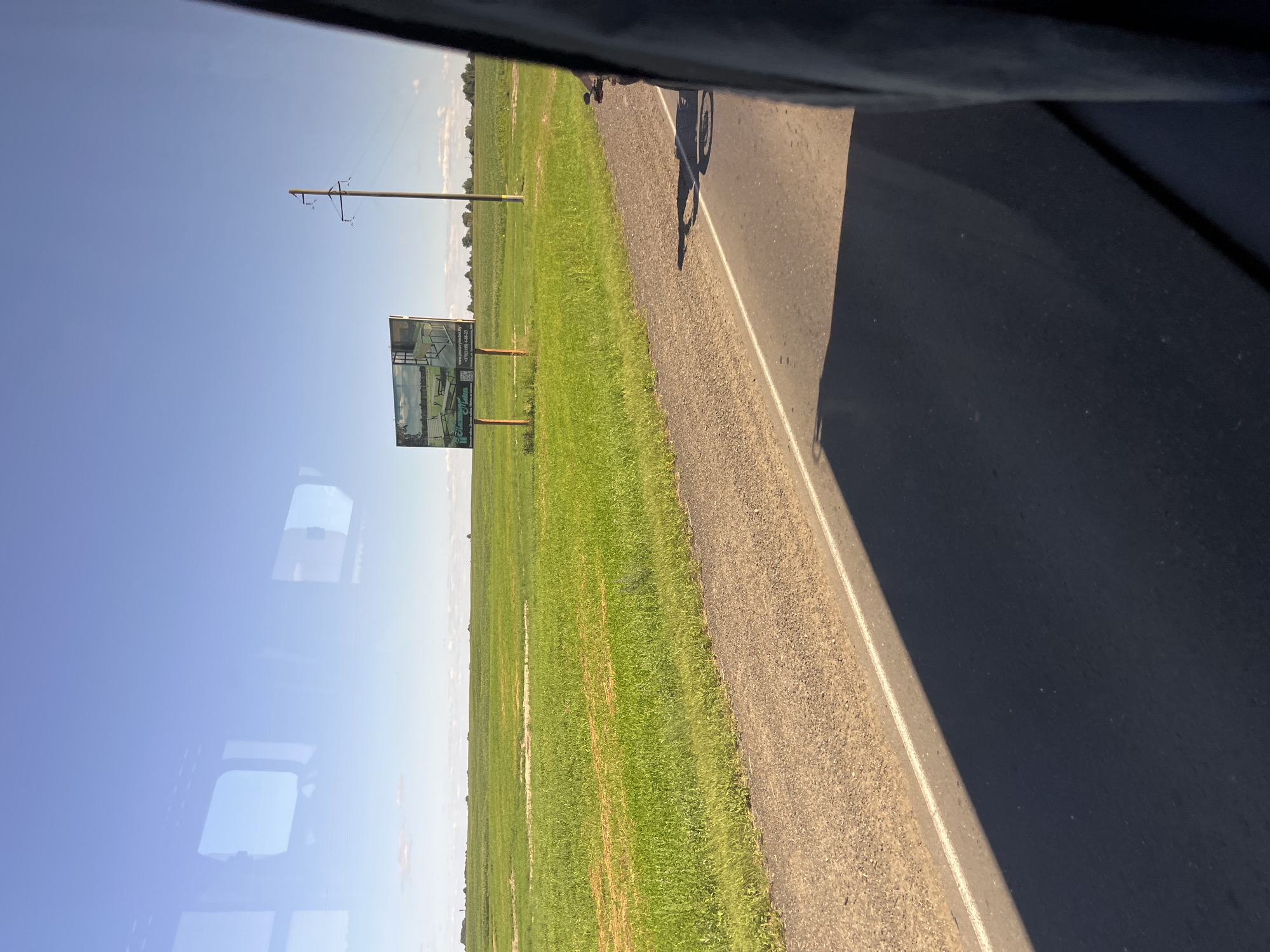 The image depicts a roadside scene in the city of Pastavy, Belarus, on a clear, sunny day. A paved road with a white line runs horizontally across the foreground, casting a long shadow from an unseen object. To the left of the road, a vast, green field stretches towards the horizon, dotted with patches of yellowing vegetation. In the distance, a billboard stands prominently in the field, displaying an advertisement with images and text, including a website address and phone number. A solitary utility pole with wires is visible to the right of the billboard, further contributing to the rural landscape. The sky is a brilliant blue, with faint reflections of what appear to be window frames visible in the upper portion of the image, suggesting the photo was taken from inside a vehicle. The overall scene conveys a sense of open space and agricultural land characteristic of the Pastavy region.