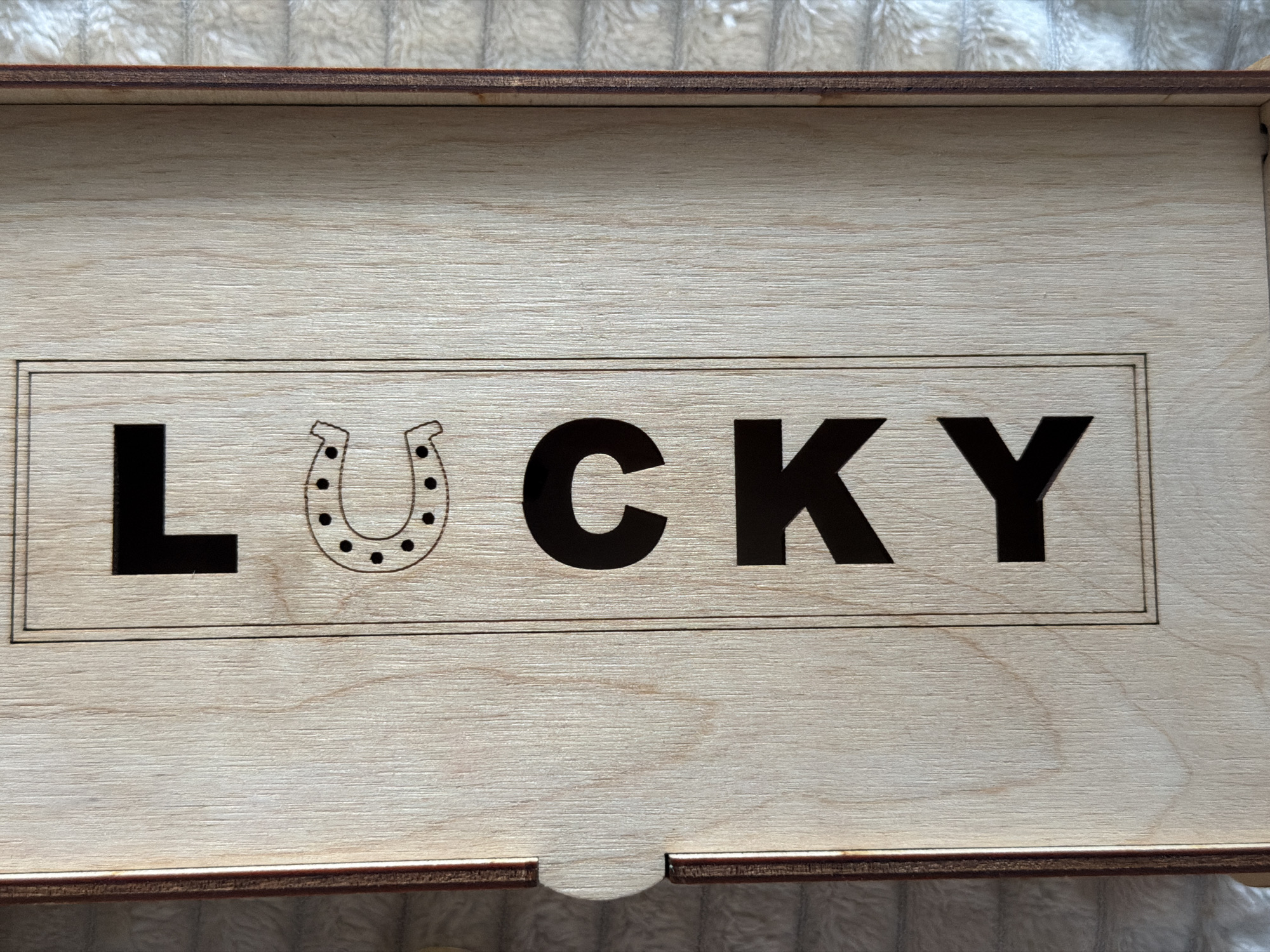The image shows a close-up view of a wooden box with the word "LUCKY" inscribed vertically on its surface. The letters are bold and black, with a horseshoe symbol placed between the 'C' and the second 'K'. The box appears to be made of light-colored wood with a visible grain pattern. The wood has a smooth finish, and the laser-cut letters and symbol are well-defined. The box has a slightly raised edge along the left side, and a small cutout detail is visible near the top left corner. The background of the image consists of a textured, light-colored surface, possibly a blanket or fabric. There is no information available about the location of this object in Kyiv, Ukraine.