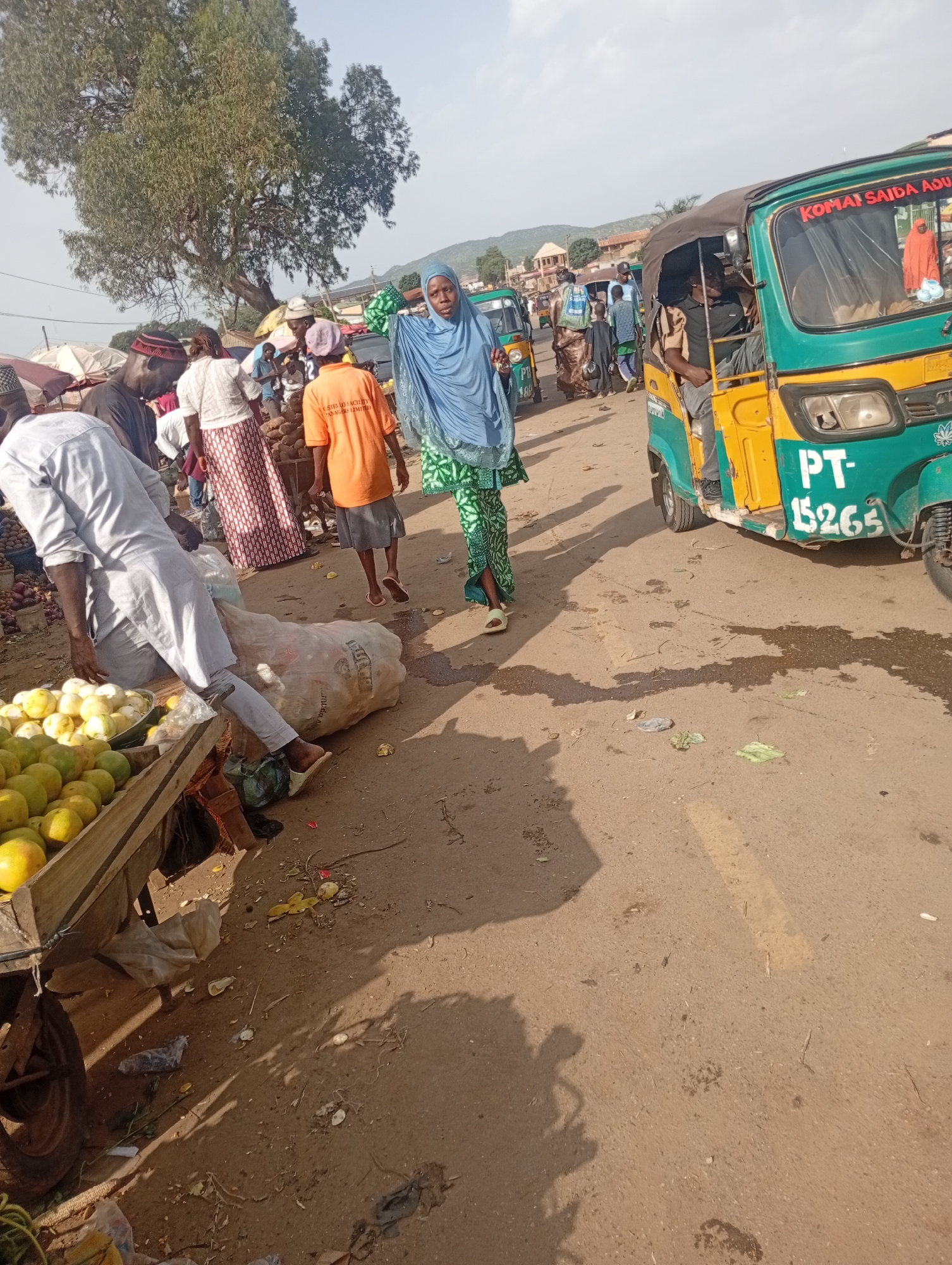 The image depicts a busy street market scene in Rafin Par, Nigeria. In the foreground, a man in a light-colored traditional garment is bent over, leaning on a cart filled with yellow fruits. To his left, more yellow fruits are piled in a wooden crate. A large, white, woven bag lies on the ground next to the man.
In the middle ground, a woman wearing a blue hijab and a green patterned dress is walking. Behind her, an orange-shirted vendor stands next to a pile of yams. Several other people are visible in the background, some wearing traditional attire. A green and yellow auto-rickshaw, marked with "PT-15265" and the words "KOMAI SAIDA ADU", is parked on the right side of the road. More auto-rickshaws and market stalls are present further in the background.
The environment is an unpaved road, with some areas appearing wet. Debris, including plastic bags and discarded food items, is scattered on the ground. A large tree with dense foliage stands in the upper left portion of the image, casting shadows. The sky is bright and overcast. The overall impression is one of a lively and active marketplace.