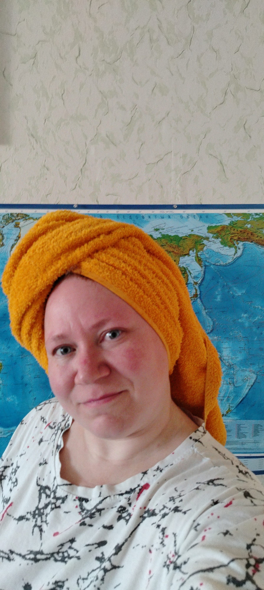 A woman with fair skin and a yellow towel wrapped around her head is looking at the camera. She has short, light-colored hair and appears to be wearing a white t-shirt with a black and red splatter-like design. The background includes a wall with a light beige patterned wallpaper on the top and a map of the world below.