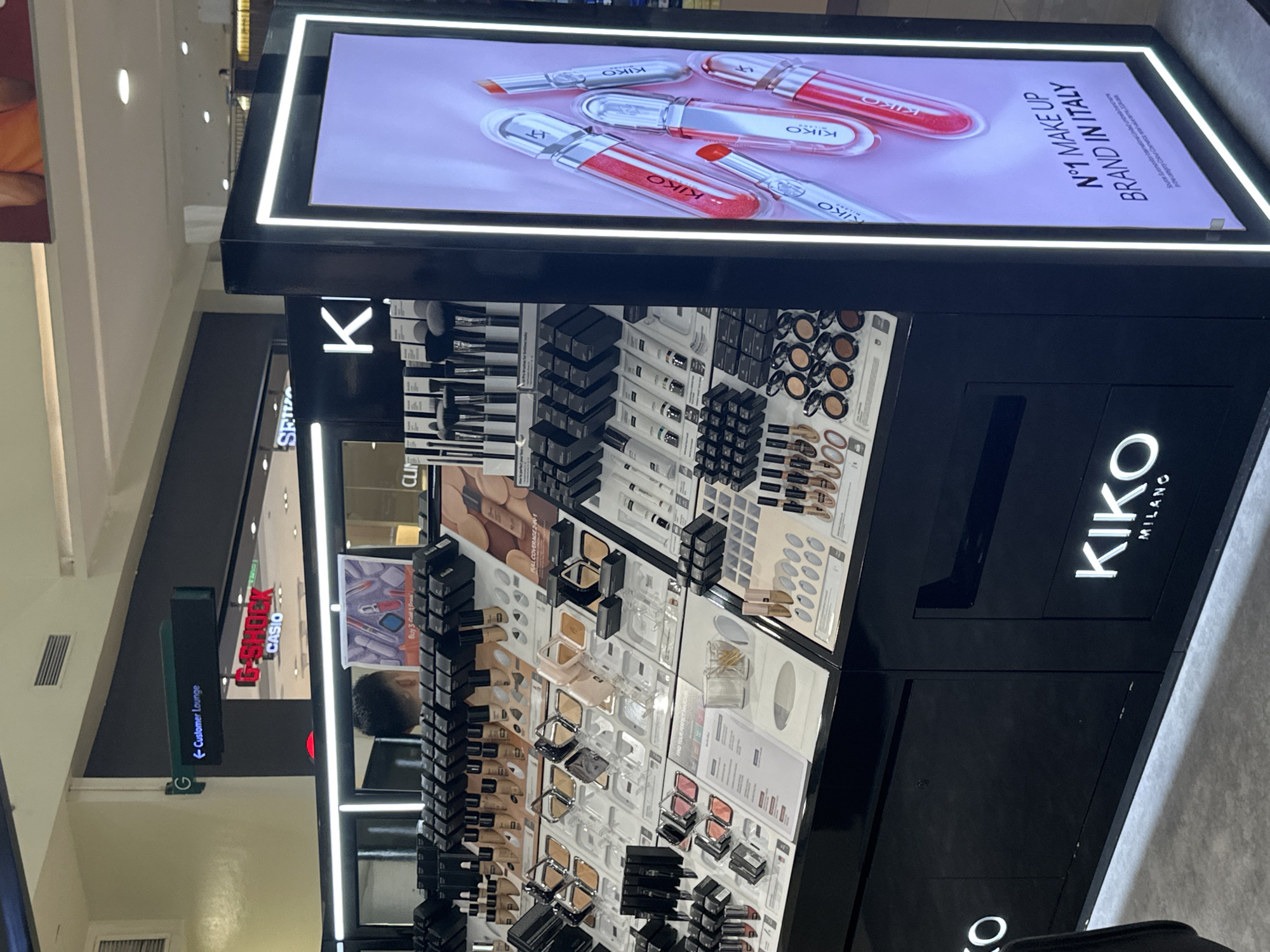 The image depicts a Kiko Milano makeup store display, likely situated within a shopping mall in Davao City, Philippines. The foreground is dominated by a sleek, black display counter showcasing a wide array of Kiko Milano beauty products, including foundations, lipsticks, and eyeshadows, arranged in clear plastic holders. The products are meticulously organized, with numerous items in their original packaging, suggesting a retail environment.
Dominating the right side of the frame is a large, illuminated digital screen displaying Kiko Milano branding and product imagery. The screen features an advertisement for "N°1 MAKE UP BRAND IN ITALY," accompanied by a close-up of several Kiko Milano lip glosses and lipsticks. The lighting from the screen casts a glow on the surrounding display.
In the background, hints of the mall environment are visible. A green directional sign with white lettering points to "Customer Lounge." Further back, store facades with logos like "G-SHOCK" and "CASIO" suggest the presence of other retail outlets within the mall. The ceiling features typical mall lighting and ventilation systems.
A person, likely a customer, is partially visible in the middle ground, viewed from the back, with their head and shoulders in frame. They appear to be looking at the makeup products on display. The floor is a light-colored, tiled surface, common in commercial establishments. The overall impression is a well-lit, modern retail space dedicated to cosmetic products.