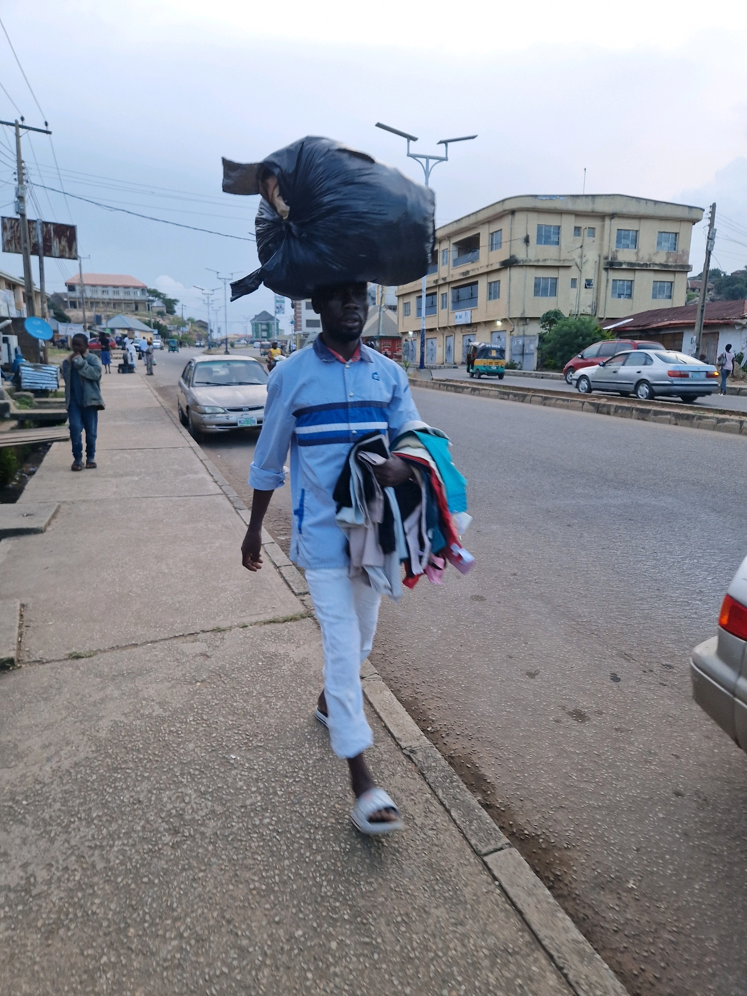 A Black man walks along a concrete sidewalk in Kabong, Nigeria, carrying a large black trash bag balanced on his head and a bundle of colorful clothing in his arms. He is wearing a light blue and white striped long-sleeved shirt and white pants, with white sandals on his feet. The sidewalk is bordered by a road with light traffic, including cars, a minivan, and a three-wheeled auto-rickshaw. Buildings with multiple stories line the road in the background, and utility poles with wires crisscross the sky. The weather appears overcast, with a hazy sky. Another person, a child, walks on the sidewalk further ahead to the left of the main subject.