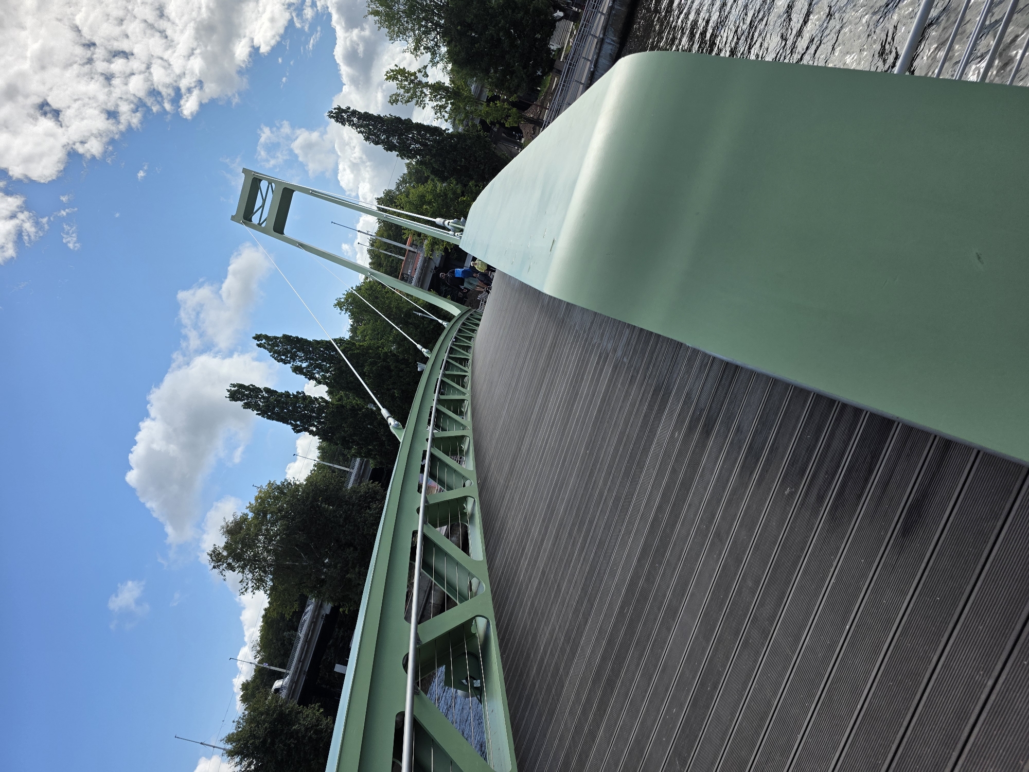 The image shows a modern, light green pedestrian bridge made of metal and wood. The bridge curves to the right, with a dark grey wooden deck. The bridge structure is intricate, with triangular supports and thin metal cables. In the background, there are lush green trees and a clear blue sky with fluffy white clouds. There are a few people visible on the bridge, including one person wearing a blue shirt and shorts, and another wearing a yellow shirt. The bridge spans over a body of water, which appears to be a river or canal, with sunlight reflecting off the surface. In the distance, there's another elevated bridge or overpass, also with trees lining the sides. The overall scene suggests a park or recreational area within a city. This bridge is known as the "King's Bridge" or "Konigsbrücke" and is located in Kaliningrad, Russia, crossing the Pregolya River.