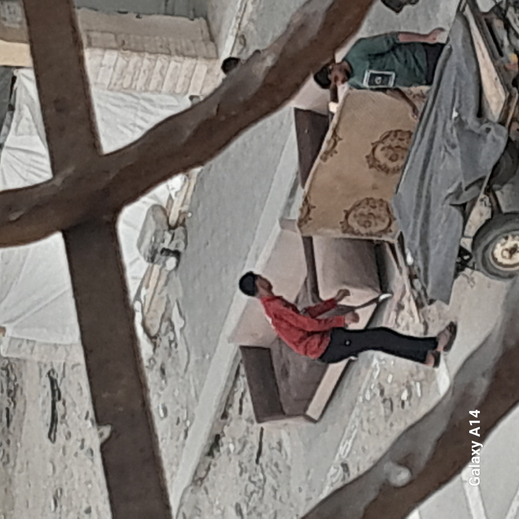 The image shows an outdoor scene in a city. A person in a red shirt and black pants is standing on a concrete structure. In the background, there is another person in a green shirt lying on a mattress. There are also various objects such as a gray cloth, a wheel, and furniture visible. A tree branch is in the foreground, partially obscuring the view.