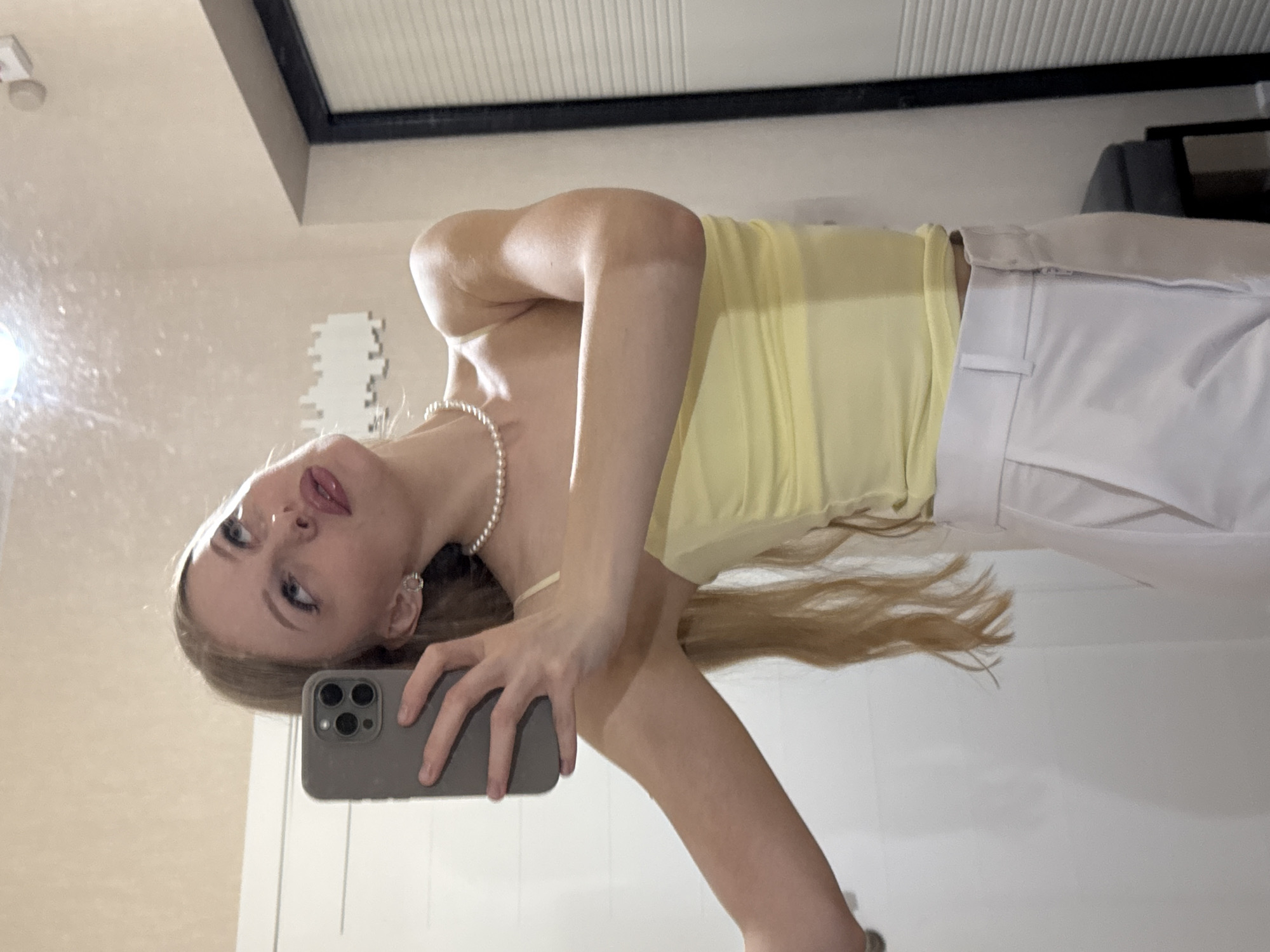 The image is a vertically oriented, slightly blurry selfie taken in a room. A young woman with long blonde hair is the sole subject, and she is positioned in the lower half of the frame, leaning her head back and looking towards the camera. She is wearing a light yellow strapless top and white pants, with a pearl necklace around her neck and small pearl earrings. She is holding a grey smartphone in her right hand, which is angled to capture the reflection in what appears to be a mirror. Her lips are slightly parted, and she has visible eye makeup. The background is out of focus but suggests an indoor environment with cream-colored walls and some decorative elements, possibly a framed picture or wall art. The lighting appears to be artificial, casting soft shadows. The overall mood conveyed is casual and self-aware. No text is visible in the image.