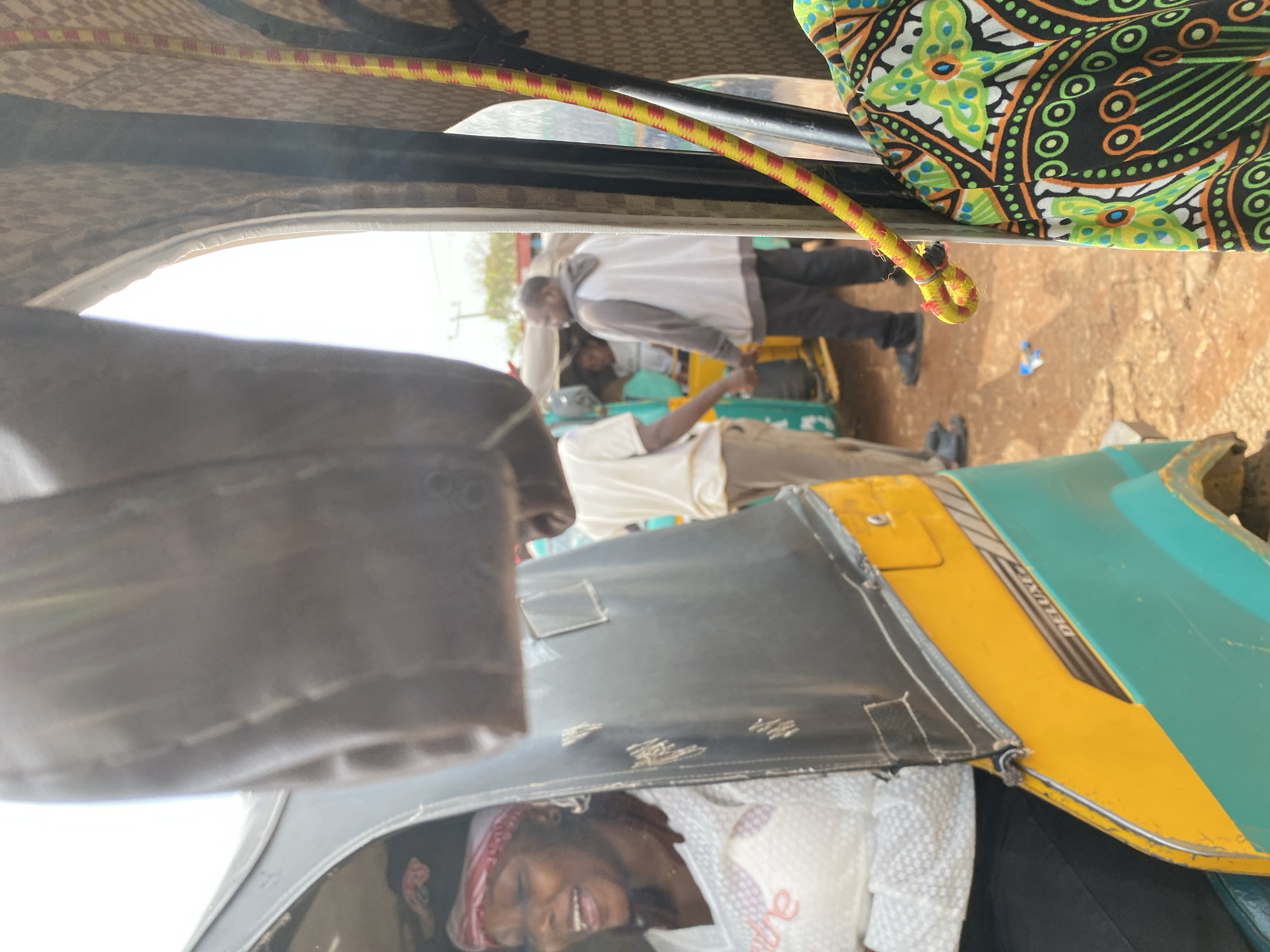 The image captures an interior view of a tuk-tuk, a three-wheeled motor vehicle commonly used as a taxi. The perspective is from inside the vehicle, looking forward and slightly upward.
**Subjects:**
In the foreground, a woman with braided hair and wearing a red and white headscarf is visible, smiling. She is partly obscured by the tuk-tuk's interior. In the middle ground, two men are standing outside the tuk-tuk. One man wears a white and gray hoodie and dark pants. Another man, wearing a light-colored t-shirt and pants, is also visible. The tuk-tuk itself is a prominent object, with its yellow and teal bodywork clearly seen, along with a black dashboard. A colorful fabric with a black, green, orange, and white patterned design is draped over the top interior of the tuk-tuk. A yellow and red elastic bungee cord runs across the top of the frame.
**Activity or Event:**
The scene appears to be a moment of rest or transition, possibly at a stop. The people are not actively engaged in a specific task, but their presence suggests a journey or waiting for passengers.
**Notable Details:**
The lighting suggests it is daytime, with bright sunlight. The ground appears to be dusty or dirt road, hinting at a less developed area. The presence of the tuk-tuk and the style of clothing worn by the individuals suggest a location in Africa, and specifically Jos, Nigeria, as per the context. The woman in the foreground has a joyful expression.
**Visible Text:**
The word "DELUXE" is visible in white lettering on a black stripe on the yellow part of the tuk-tuk's body.