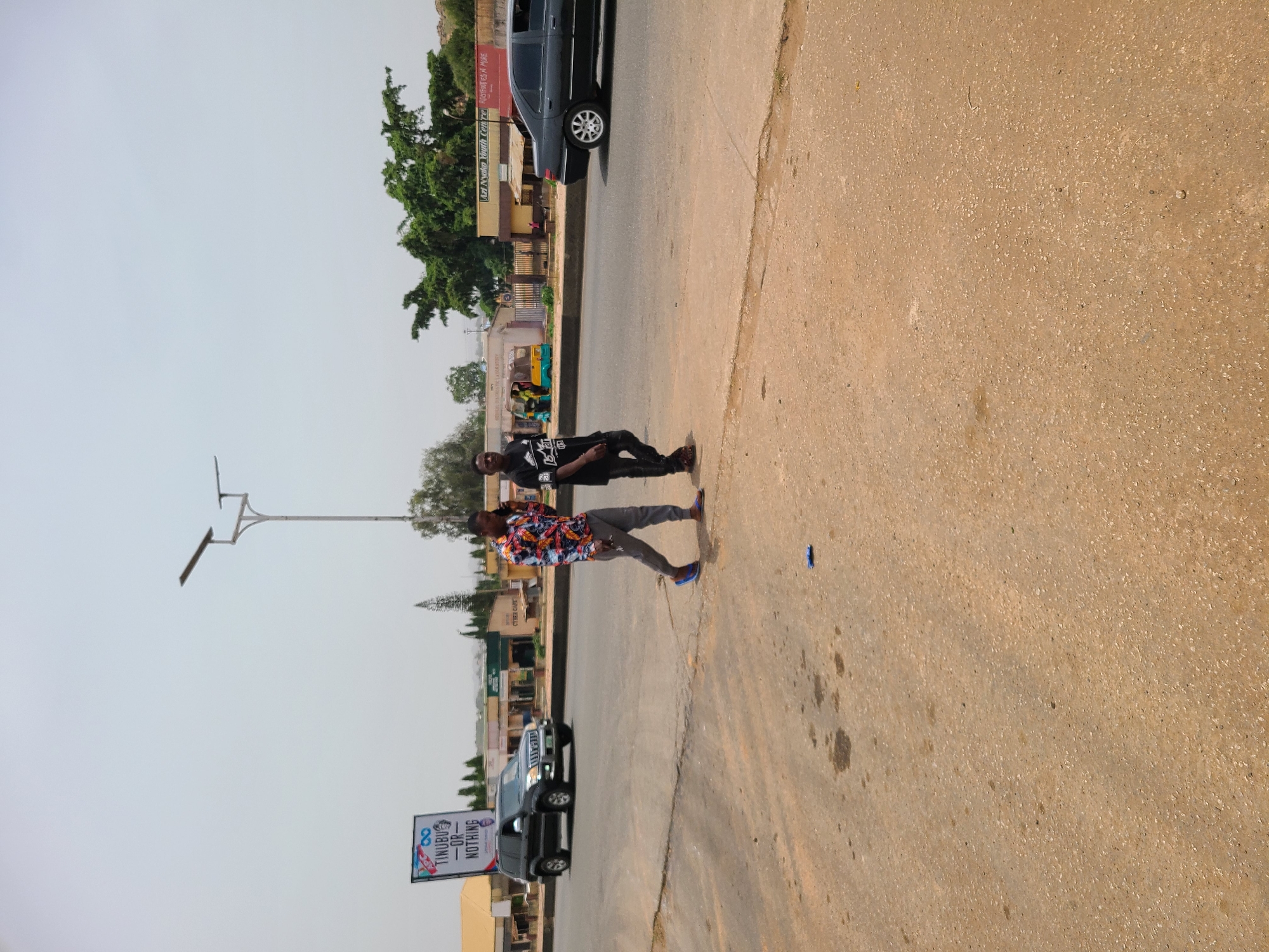 The correctly oriented image captures a daytime scene on a street in an urban or semi-urban area. Two young men are seen on the unpaved shoulder beside a road. The man in the foreground wears a colorful, long-sleeved patterned shirt, grey pants, and blue shoes. Behind him, another man wears a black t-shirt with white text and black pants, paired with brown sandals. They appear to be walking or casually standing.
On the asphalt road to their left, a dark-colored car, possibly an SUV or minivan, is visible. Further down the street, another black SUV is parked near a large billboard. The billboard prominently displays the text "TINUBU -OR- NOTHING," indicating a political campaign, with faint text "REVOLUTION NOW" also visible.
Buildings line the left side of the road, including one with a red sign reading "A.H. Nuhu Youth Centre" and another with a green and blue sign for a "Cyber Cafe." Trees and foliage are interspersed among the buildings. A light pole equipped with a solar panel stands on the left. A three-wheeled vehicle, resembling a keke napep, is also parked further down the road. The sky is clear, suggesting bright, sunny weather. The ground where the men are standing is a light brown, dusty surface, contrasting with the darker paved road. This scene depicts everyday activity on a street in Jos, Nigeria.