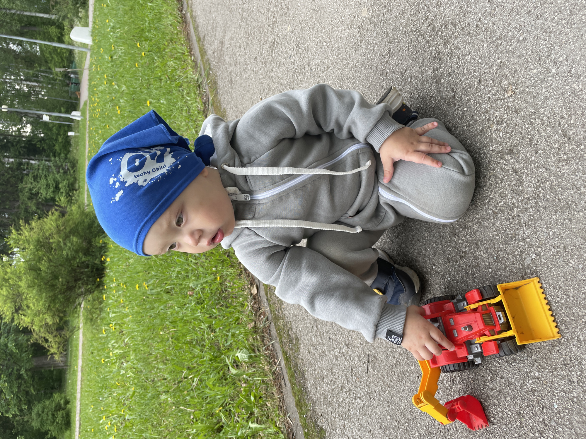 A young child, likely a toddler, is depicted outdoors, kneeling or crouching on a grey asphalt surface, engrossed in playing with a toy. The child wears a blue beanie with white text that reads "lucky Child" and a stylized graphic, along with a grey hooded tracksuit with a white drawstring and zipper. Their gaze is directed downwards, focused on the toy excavator/loader in front of them, with their right hand resting on its body.
The toy is predominantly red and yellow, featuring a large yellow front bucket and a red backhoe attachment. The setting includes a lush green grassy area with scattered yellow dandelions bordering the paved surface to the child's left and in the background. Beyond the grass, there is a dense line of green trees and bushes, and some indistinct white, vertical structures are visible further back.
The activity shows the child actively engaged in play with the construction toy. It is daytime with soft, even lighting, suggesting mild weather, possibly spring or summer. Visible text includes "lucky Child" on the beanie and "FUNNY RIDE 2018" on a small black tag on the left sleeve cuff of the tracksuit, as well as on an internal tag near the zipper. The image captures a moment of focused outdoor play. The provided location context is Perm, Russia.