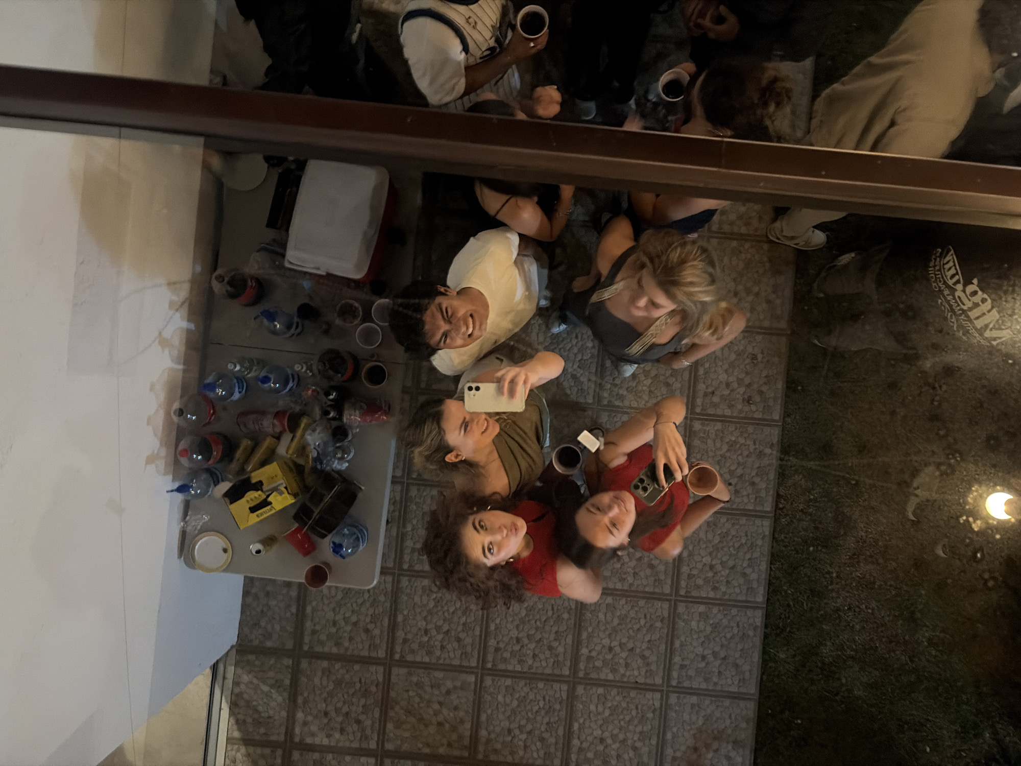 A high-angle shot, possibly taken through a window or from an upper level, captures a group of people in what appears to be an outdoor gathering. Four individuals in the foreground are looking up, two holding smartphones and taking selfies. They are standing on a tiled surface, adjacent to a grassy area illuminated by a light source. Another group of people is visible in the background, some holding cups. A table is partially visible on the left side, holding bottled water and other items. The overall setting suggests a casual social event. The location is Unknown, Unknown.