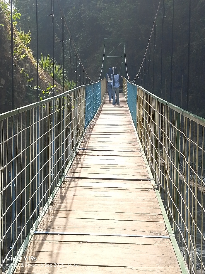 The image displays a long suspension bridge in Sukadana, Indonesia, viewed lengthwise from one end. The bridge features a walkway constructed of horizontal wooden planks. Its side barriers consist of metal mesh fencing, with structural elements painted in alternating yellow and blue, topped by green horizontal rails. Vertical and diagonal steel cables descend from main suspension lines to support the deck. Two individuals are observed walking away from the camera on the bridge's walkway towards the distant end. The person on the left wears a light-colored long-sleeved top, dark trousers, and a dark head covering. The person on the right wears a blue long-sleeved top, light-colored trousers, and a dark head covering. Dense green vegetation covers the steep bank on the left side of the bridge and forms a thick, dark background beyond. Bright sunlight from the left casts distinct, elongated shadows across the wooden planks and from the bridge's structural elements. The environment indicates clear weather.