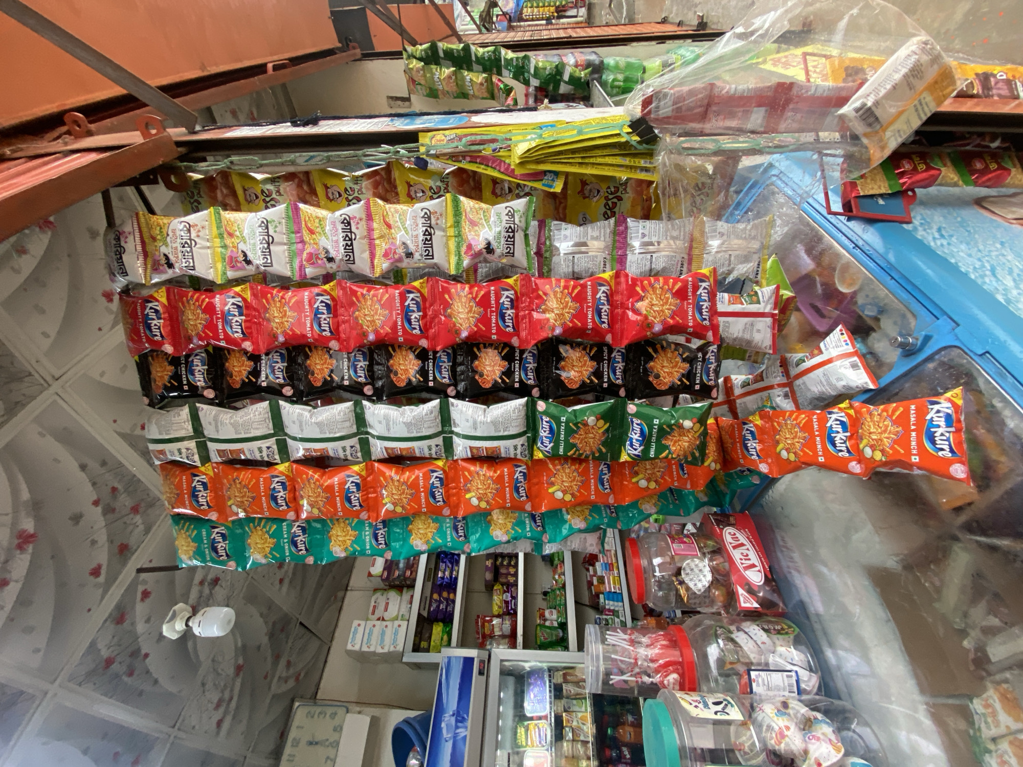 The image captures the interior of a small roadside shop, likely located in Dhaka, Bangladesh. The shop’s walls and ceiling are lined with various snack items, primarily packets of Kurkure in various flavors such as "Naughty Tomato", "Spicy Chicken", "Masala Munch", and "Cream & Onion". There are also packets of other snacks with Bengali script on the labels. The products are densely packed, suspended from the ceiling and displayed on shelves. Below the hanging snacks, shelves are stocked with additional items, including what appear to be medicines and other goods in small packages. A small refrigerator stands near the bottom of the frame, filled with items. There are also jars and plastic containers filled with candies and other items. The lighting in the shop appears to be provided by a light bulb and natural light filtering in. The scene suggests a typical small business providing convenience goods in an urban area.