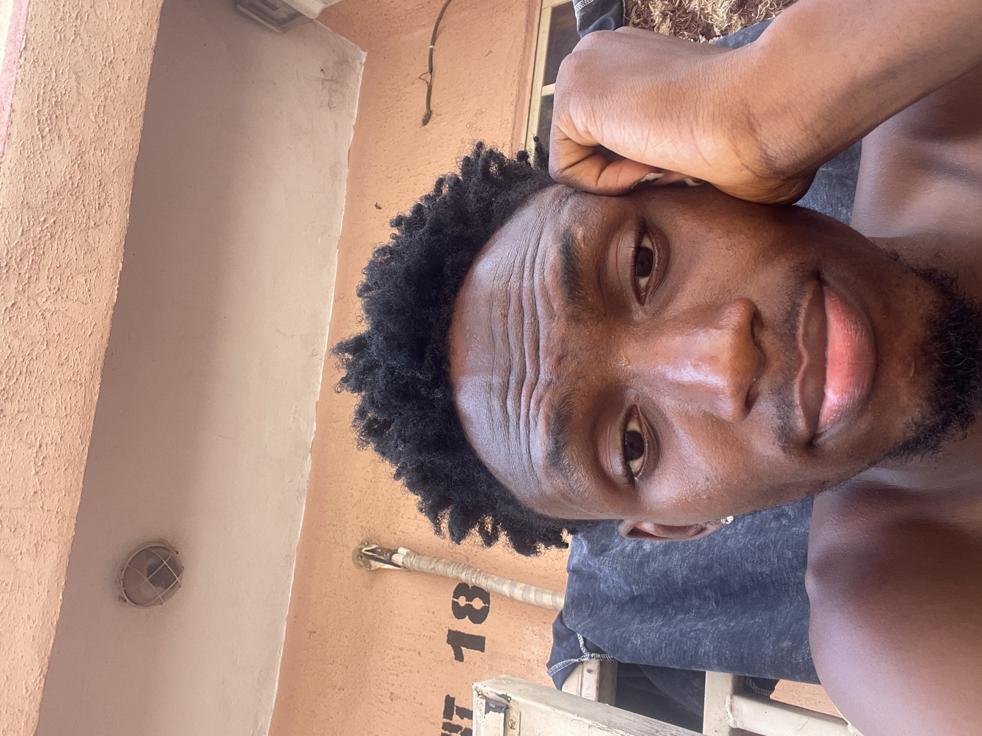 A close-up, angled selfie captures a young man with dark skin and dark, curly hair looking directly at the camera. His head is tilted, and his left forearm and hand rest above his temple. Wrinkles are visible on his forehead, suggesting a thoughtful or pensive expression, though his lips are slightly parted. He has stubble on his chin and upper lip. He appears to be wearing a dark, possibly grey, casual shirt.
The background features textured walls in varying shades of peach, orange, and light beige, suggesting an outdoor or semi-outdoor setting in Lokoja, Nigeria, likely during daylight hours given the clear lighting and reflections on his skin. A semi-circular light fixture with a grid pattern is visible on the light-colored wall. Below it, a horizontally positioned, wrapped pipe-like object is attached to the wall. Prominently displayed beneath this pipe are the black numbers "18" and a partial letter, possibly "F" or "T", on the peach-colored wall. Another thin, dark wire or pipe runs vertically higher up on the left wall. The overall scene conveys a casual, personal moment captured in a warm, possibly humid environment.