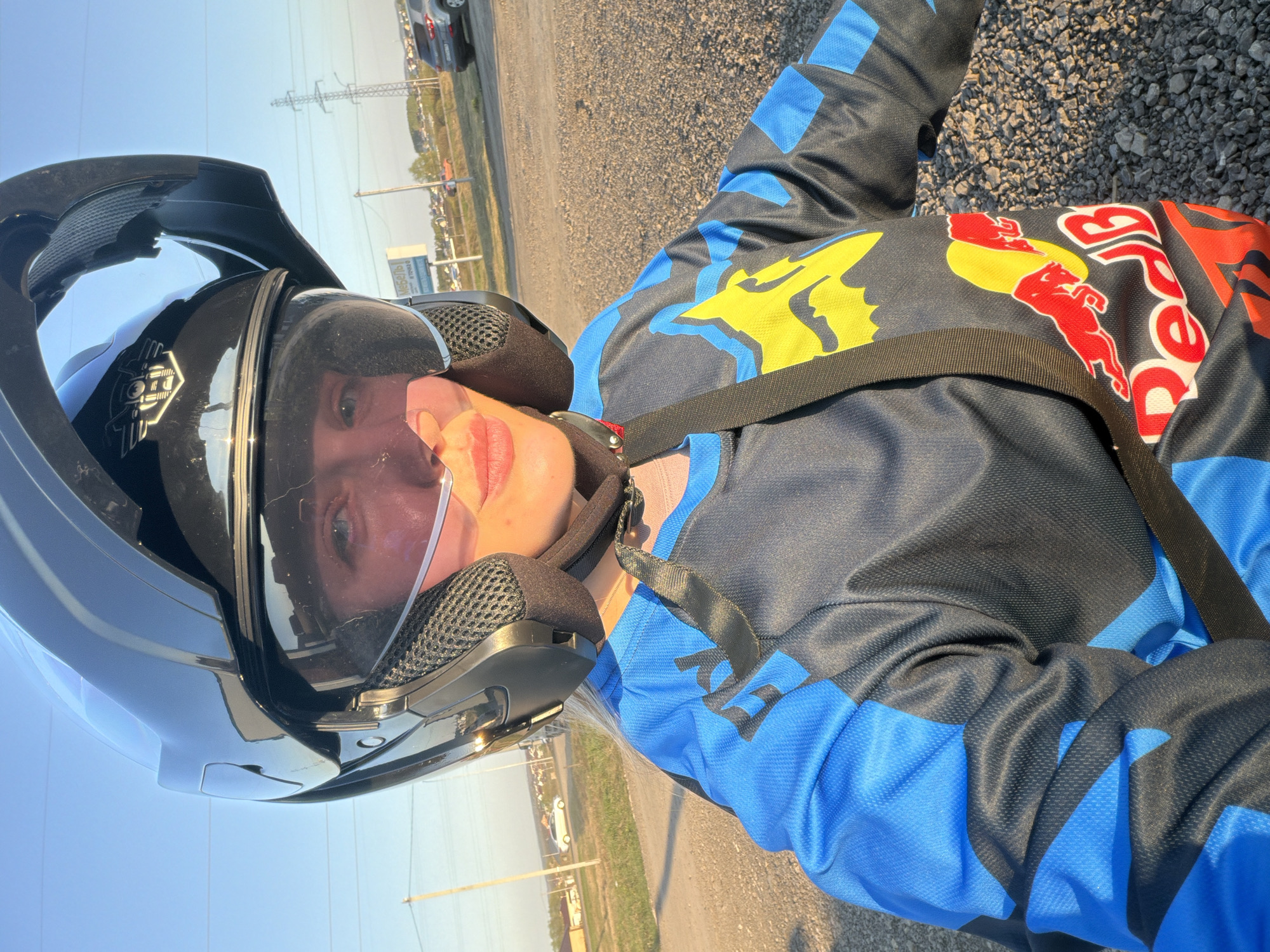 A person wearing a black motorcycle helmet and a blue and black racing-style jacket with "Red Bull" and "Fox" logos is seen in an outdoor setting. The person has a nasal cannula in their nostrils. The ground appears to be gravel or small stones. In the background, there is a road, grassy area, and utility poles. There is a building in the distance. The location may be Азьмушкино, Russia, based on the instructions.