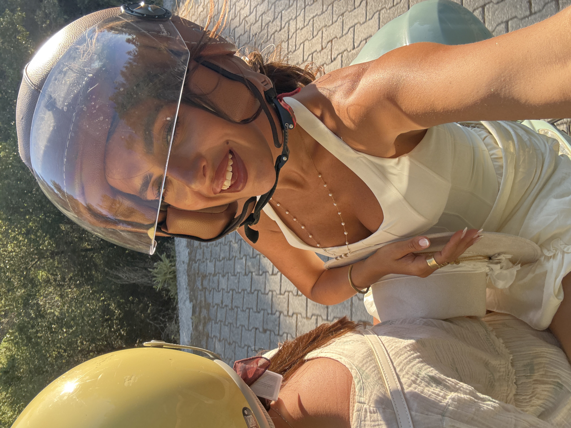 This media file captures two individuals on a scooter, presumably in Fethiye, Turkey. The primary subject, in the foreground, is a woman wearing a brown helmet with a clear visor and a white top. She is smiling and looking towards the camera, with her mouth open in laughter. Her hair is dark brown and slightly windswept. She is wearing a delicate pearl necklace, a gold bracelet on her left wrist, and a gold ring on her right index finger. The second individual is partially visible in the lower portion of the image, wearing a yellow helmet. They appear to be the driver of the scooter, as evidenced by the presence of the yellow helmet.
The scene is outdoors, with a cobblestone-like paved surface visible behind the woman. Green foliage and trees are present in the background, suggesting a sunny day and a warm climate. The lighting indicates it is daytime, likely late afternoon or early morning given the warm, angled sunlight. The overall mood is cheerful and spontaneous, suggesting a recreational outing or travel experience. The presence of the helmets and the scooter indicates they are in motion or have just stopped.
Notable details include the textured leather finish of the brown helmet, the delicate pearl necklace, and the woman's manicured fingernails. The second person's attire includes a light-colored, possibly linen garment. There is no visible text in the image.