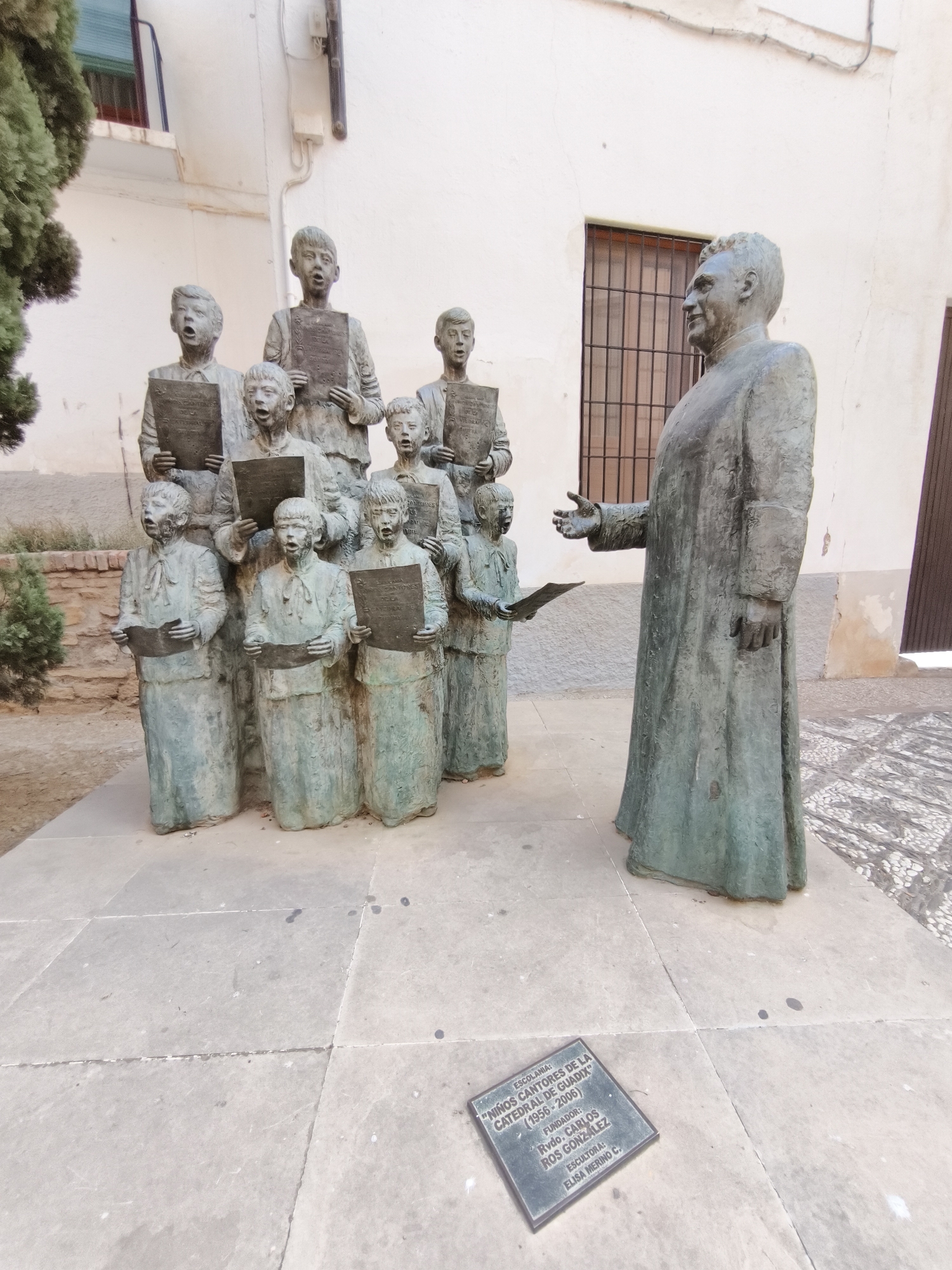 The image shows a bronze sculpture of a group of children singing, led by an adult male figure, likely a choirmaster or priest. The children are depicted holding sheets of music and have their mouths open as if singing. The adult figure stands to the right, facing the children and extending his left hand. A plaque at the base of the sculpture reads "ESCOLANIA: 'NIÑOS CANTORES DE LA CATEDRAL DE GUADIX' (1956-2006). FUNDADOR: Rvdo. CARLOS ROS GONZALEZ. ESCULTORA: ELISA MERINO C.". This indicates the sculpture is located in Guadix, Spain, and commemorates the Children's Choir of the Guadix Cathedral, founded by Carlos Ros Gonzalez and sculpted by Elisa Merino. The sculpture is situated on a paved courtyard in front of a building with white walls and barred windows. There is a tree with green foliage on the left side of the image and a dark brown door on the right. The lighting suggests it is daytime.