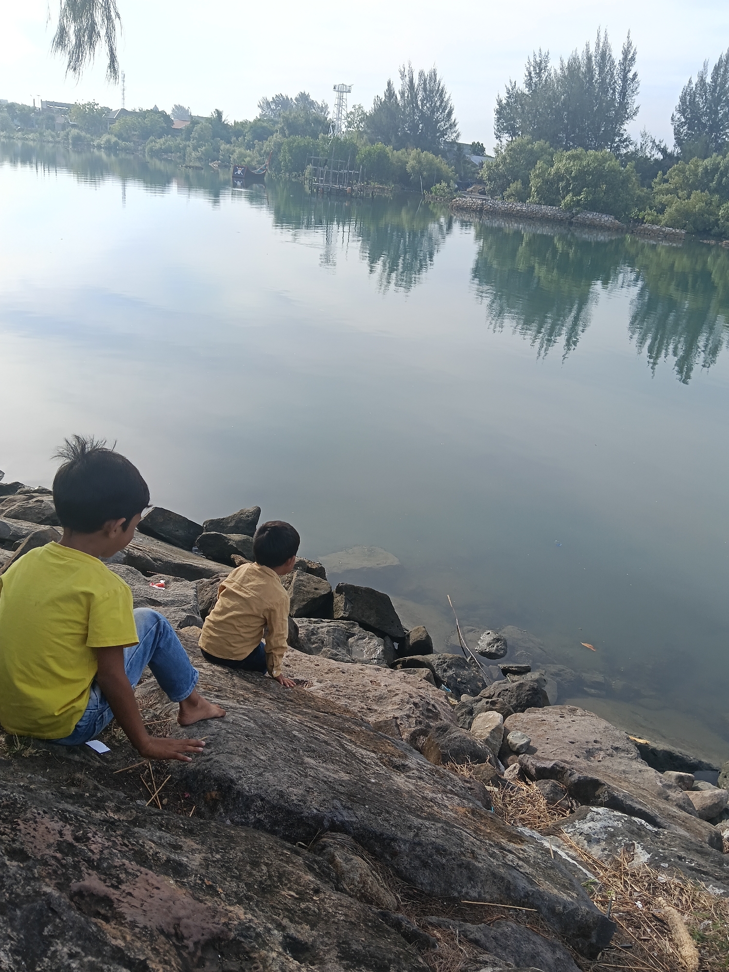 Two young boys, one in a yellow t-shirt and blue jeans, and another in a tan long-sleeved shirt, are sitting on a rocky embankment by a body of water. The boy in yellow is closer to the foreground and is turned to his left, facing the water. The boy in tan is further back and is also facing the water. The water is calm and reflects the trees and sky on the opposite bank, which is lined with vegetation and some structures, including what appear to be communication towers and buildings in the distance. The embankment is made of large, grey rocks, with some dry grass and debris scattered among them. The overall scene suggests a natural or semi-natural setting within or near Banda Aceh, Indonesia, possibly a river or coastal area.