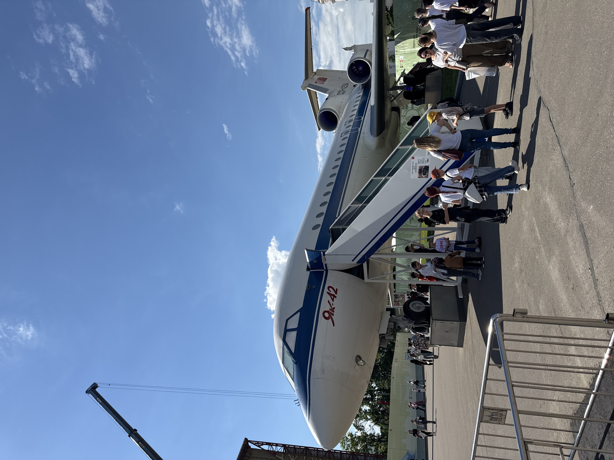The media file, rotated 90 degrees clockwise, depicts an outdoor scene under a clear blue sky with scattered white clouds, showcasing a large passenger aircraft on static display. The prominent subject is a Yakovlev Yak-42 airplane, identifiable by "ЯК-42" (Yak-42) written in red Cyrillic script on its nose and the Soviet-era registration "CCCP-42314" on its tail. The aircraft, painted in a white and blue livery, features three rear-mounted engines, characteristic of the Yak-42, and has an airstair extended to its main door, allowing visitor access.
Numerous people, dressed in casual clothing, are gathered around the aircraft and queueing on the paved ground, seemingly waiting to enter or exit the plane. Some individuals are visible on the airstair, ascending towards the fuselage. The setting appears to be an aviation exhibition or museum grounds in Khimki, Russia. In the background, a large green informational banner or wall with indistinct Cyrillic text and diagrams provides additional context to the display. A crane arm bearing the text "АКМ" is partially visible in the bottom left, suggesting nearby equipment or construction. A metal crowd control barrier stands in the foreground on the right. The bright daylight and clear weather suggest a pleasant day for the event.