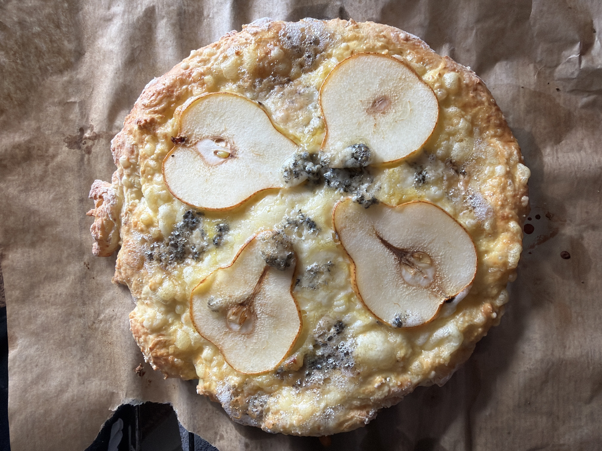 The image shows a pear and blue cheese tart on a sheet of brown parchment paper. The tart is round with a golden-brown, slightly irregular crust. Four pear slices are arranged in a circular pattern on top of the tart, with crumbles of blue cheese scattered between them. The parchment paper has wrinkles and folds, and a small portion of a dark object is visible in the lower left corner of the frame. The location is not discernible from the image.