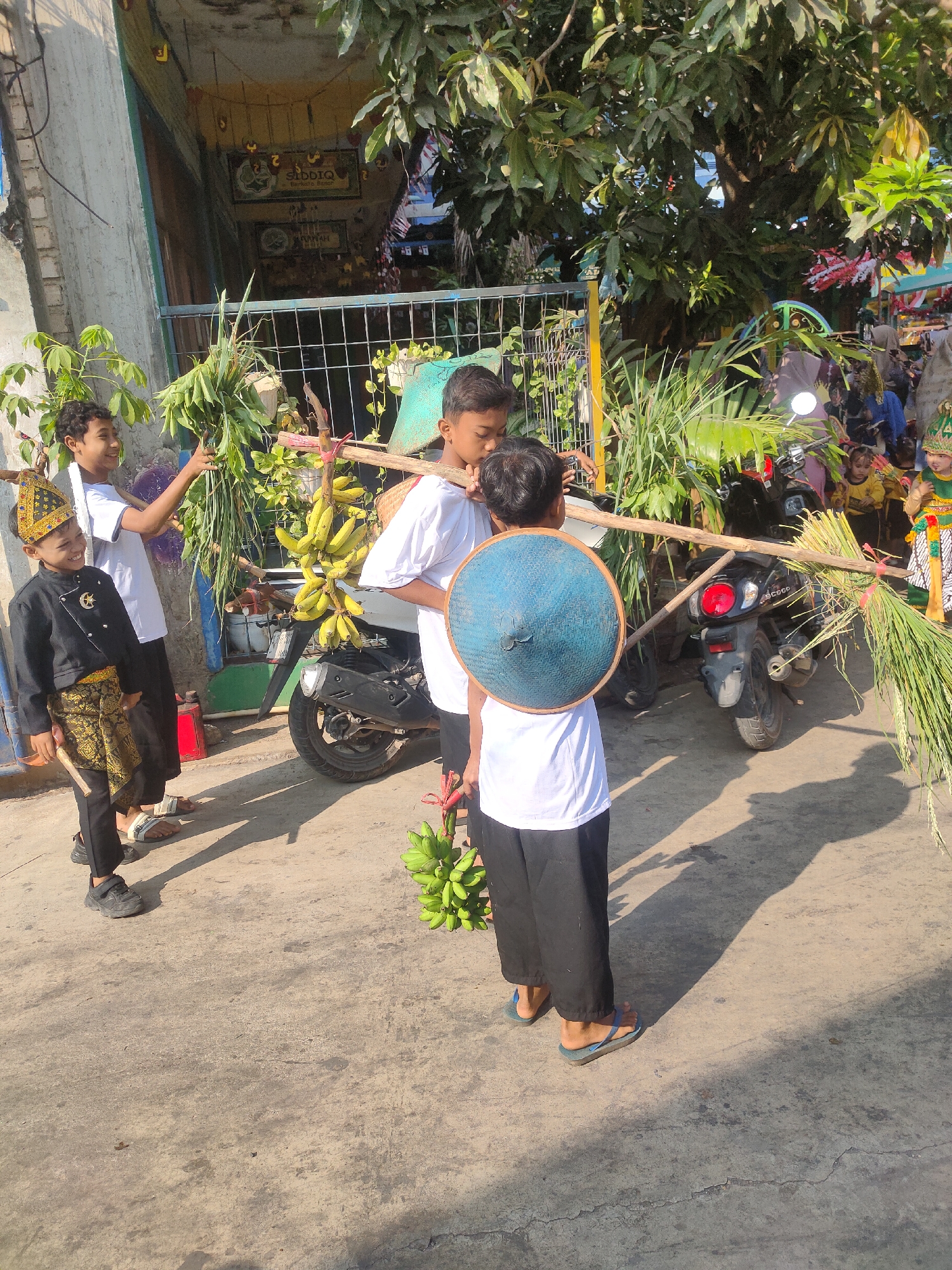 In the city of Payaman, Indonesia, on a sunny day, four young boys are captured in a candid moment, carrying offerings. The boy on the far left, adorned in a black and gold traditional outfit, smiles as he walks alongside a motorbike. He holds a bundle of green foliage in his left hand. To his right, another boy in a white t-shirt and black pants carries a long bamboo pole over his shoulders. This pole is laden with bunches of green bananas. He appears to be engaged in conversation with the boy to his right. The boy on the far right, also in a white t-shirt and black pants, wears a blue conical hat on his back. He holds the other end of the bamboo pole, which is also decorated with stalks of rice. In the background, a motorbike is parked, and a fence separates the scene from a building adorned with colorful decorations and signs, one of which reads "SIDDIQ." Other children, some in traditional attire, can be seen in the distance. The ground is paved with concrete, and shadows of the people and trees are cast across it.