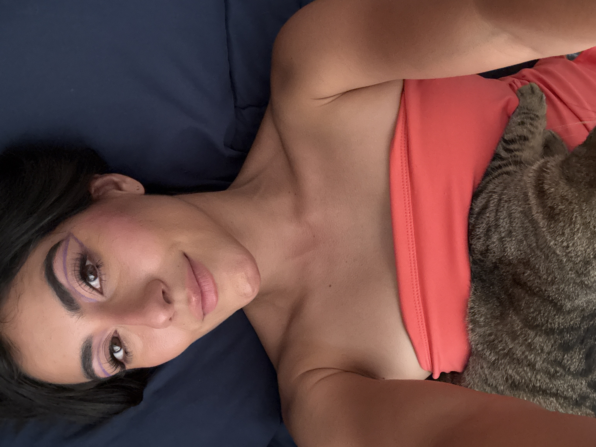 A woman with dark hair and bold, colorful eye makeup lies on her back on a dark blue fabric surface, possibly a bed. She is wearing a coral-colored top. A tabby cat is positioned on her right side, partially covering her. The location in the city of Puebla, Mexico, is not evident in the image.
