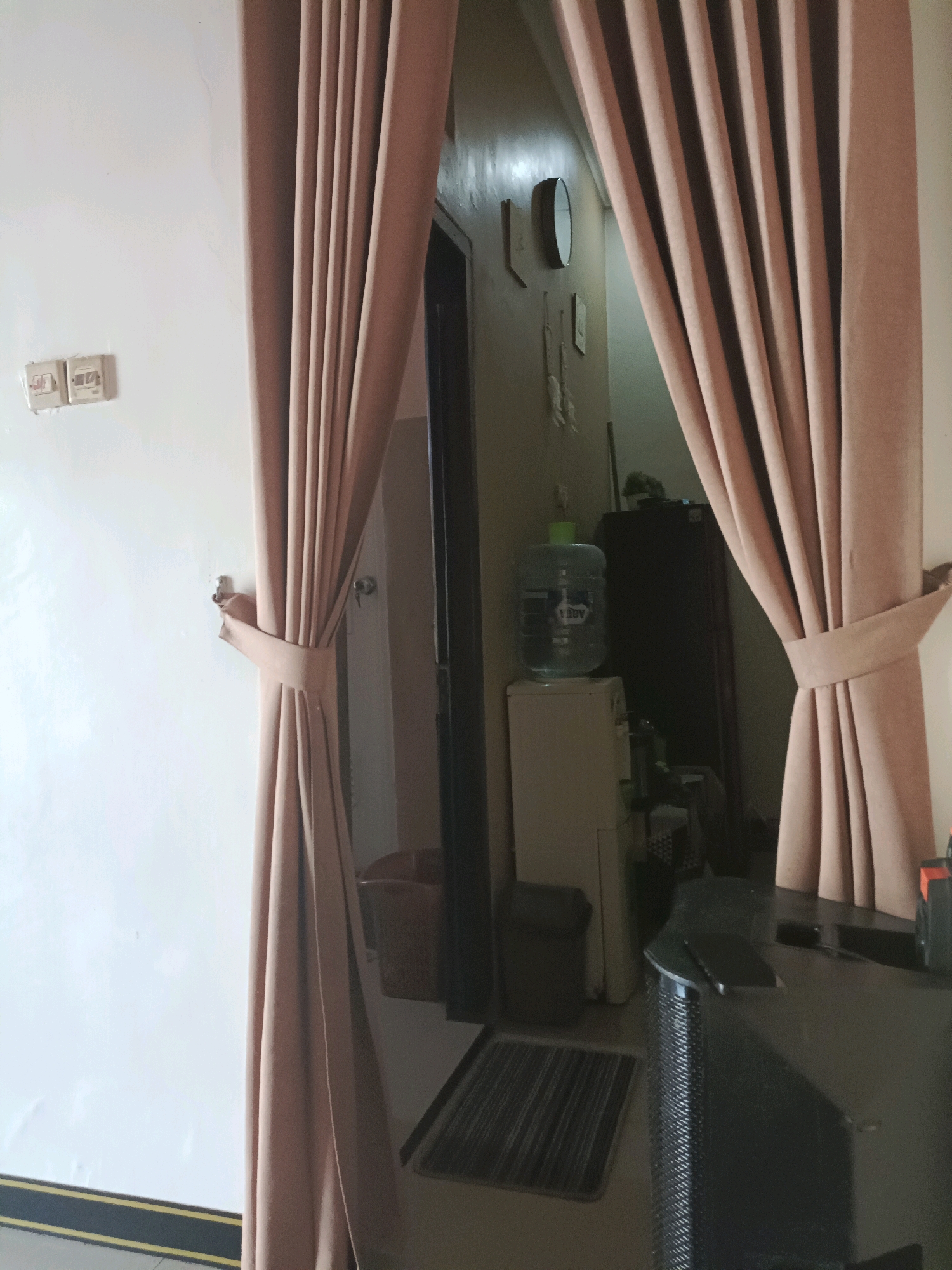 A pinkish-beige curtain is tied back on the left side of the frame, revealing a doorway leading into a hallway. The wall on the left is painted white and features two electrical outlets. The hallway beyond the curtain is dimly lit. A water dispenser with a large blue bottle is visible in the hallway, positioned on top of a small, cream-colored appliance. Further down the hallway, a dark cabinet is present, and a clock hangs on the wall. On the right side of the frame, the curtain continues to hang, partially obscuring a dark countertop with a black speaker and a black electronic device. A grey doormat is visible on the floor in the hallway, leading towards a dark entryway. The overall environment suggests an indoor residential or office space in Rancang Kencono, Indonesia.