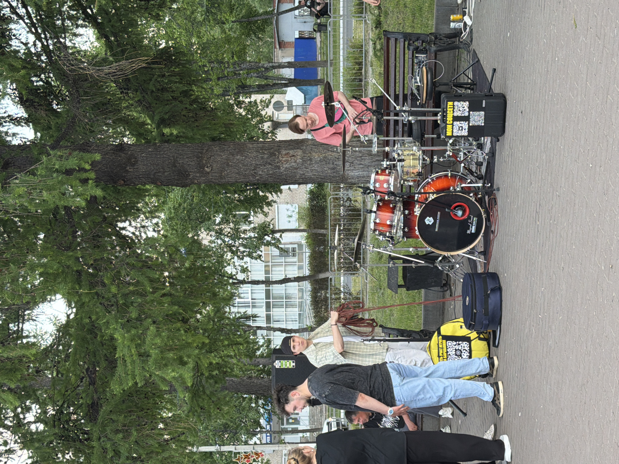 A man plays a drum set in a public space in Chaikovsky, Russia. The drummer, wearing a pink t-shirt, is positioned behind the drum set. Several individuals stand around, appearing to watch the performance. A large tree dominates the left side of the frame. Buildings and other urban elements can be seen in the background.