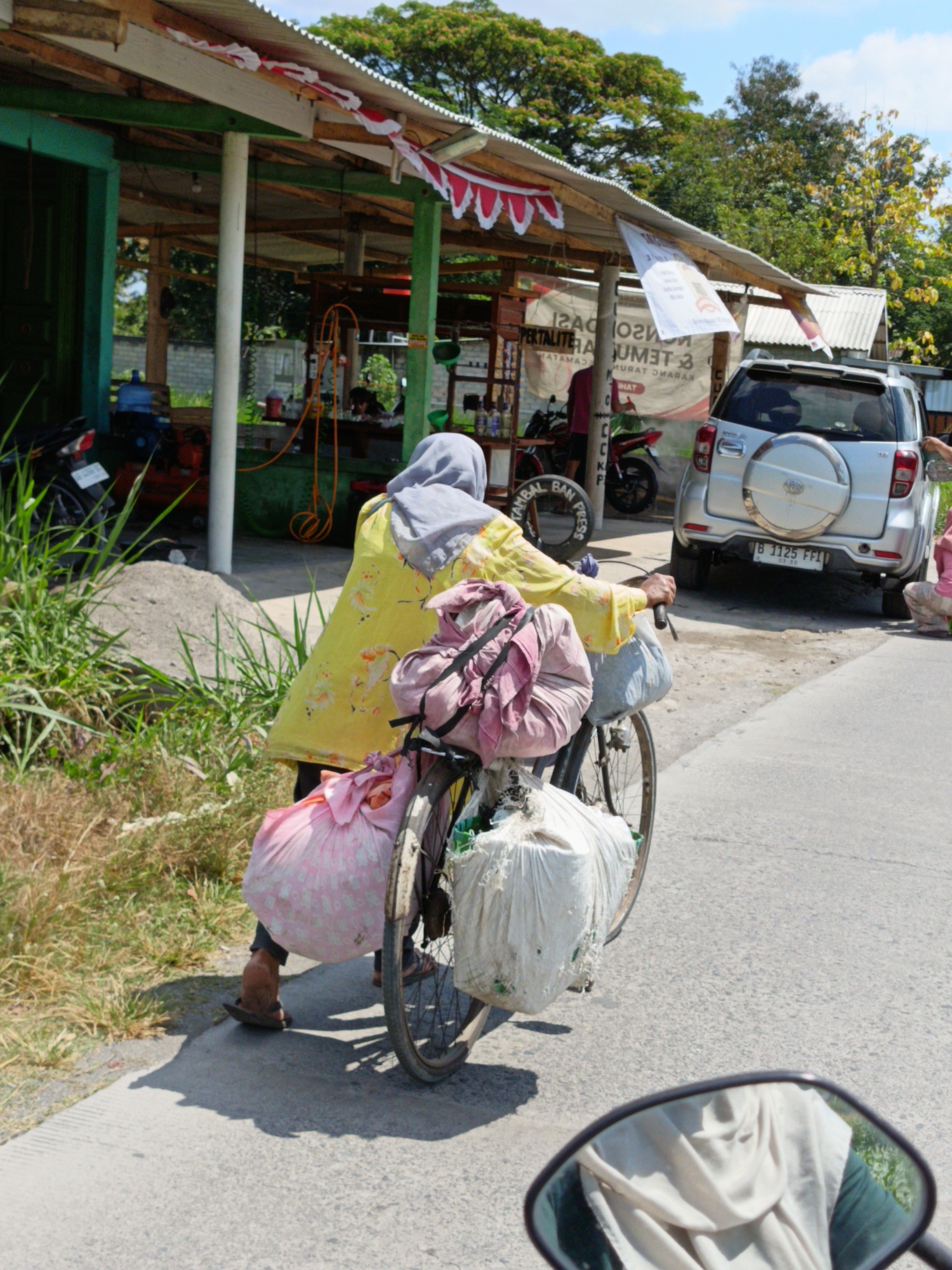In Nangsri, Indonesia, a person wearing a yellow floral patterned dress and a grey headscarf is pushing a bicycle laden with several large bags. One pink bag is attached to the front of the bicycle, and a large white bag is secured to the rear. The person's legs are visible, wearing dark pants and brown sandals. The bicycle has a black frame and a spoked rear wheel, with a partial view of the front wheel. In the background, a silver SUV with a spare tire mounted on the back is parked on the right side of the road, displaying a license plate that reads "B 1125 FFI". To the left of the road, a building with a corrugated metal roof and a red and white striped awning is visible, with several motorcycles parked nearby. The ground is paved with asphalt, and there is vegetation on the left side of the image. The sky is bright and appears to be sunny.