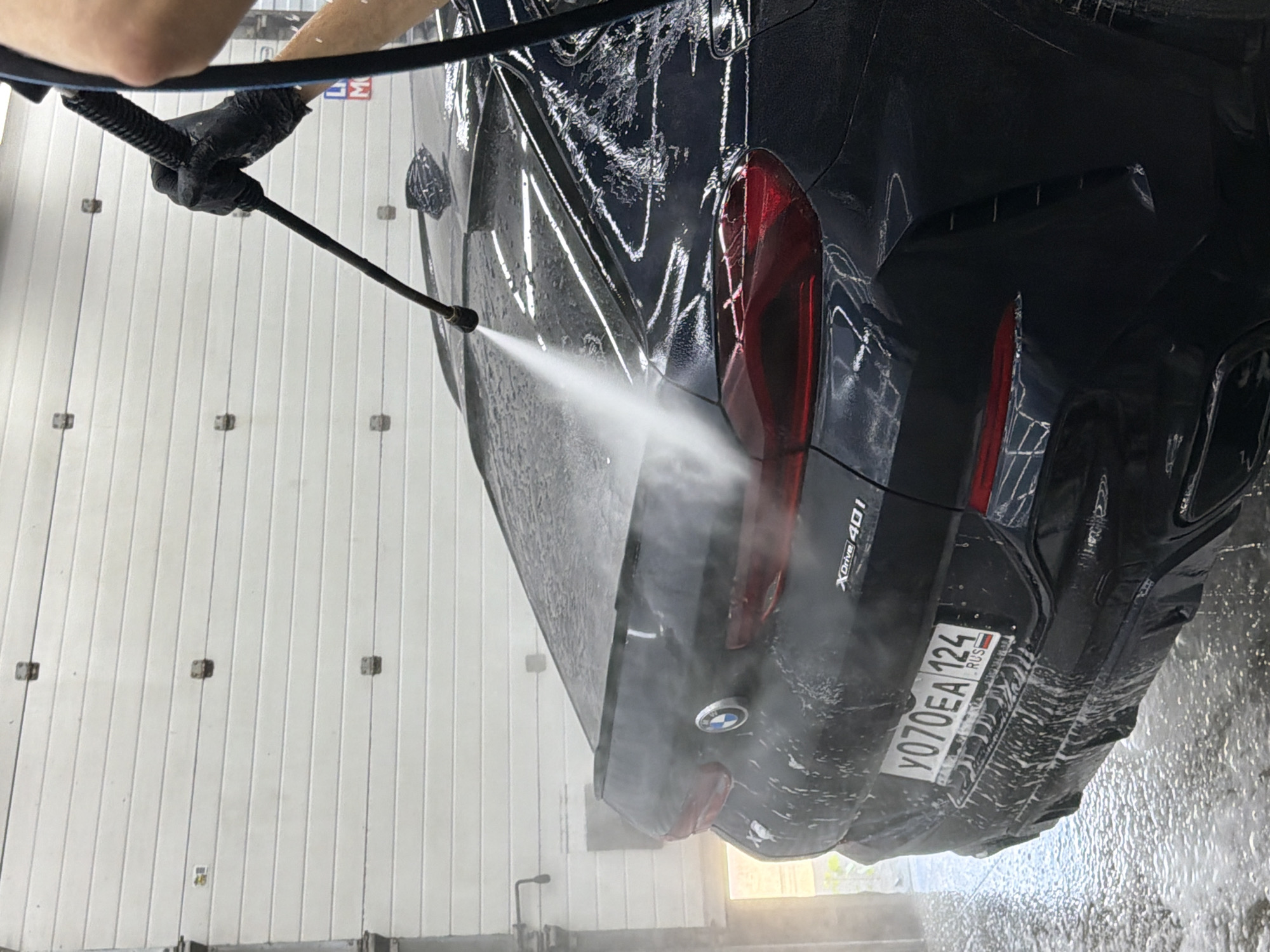The image shows a person in black gloves washing a black BMW X6 with a high-pressure water spray. The car has a Russian license plate that reads "Y070EA 124". The car is inside a garage or car wash, with a white corrugated metal door in the background. There are signs of water and soap suds on the car and the floor. The overall scene depicts a car being cleaned.