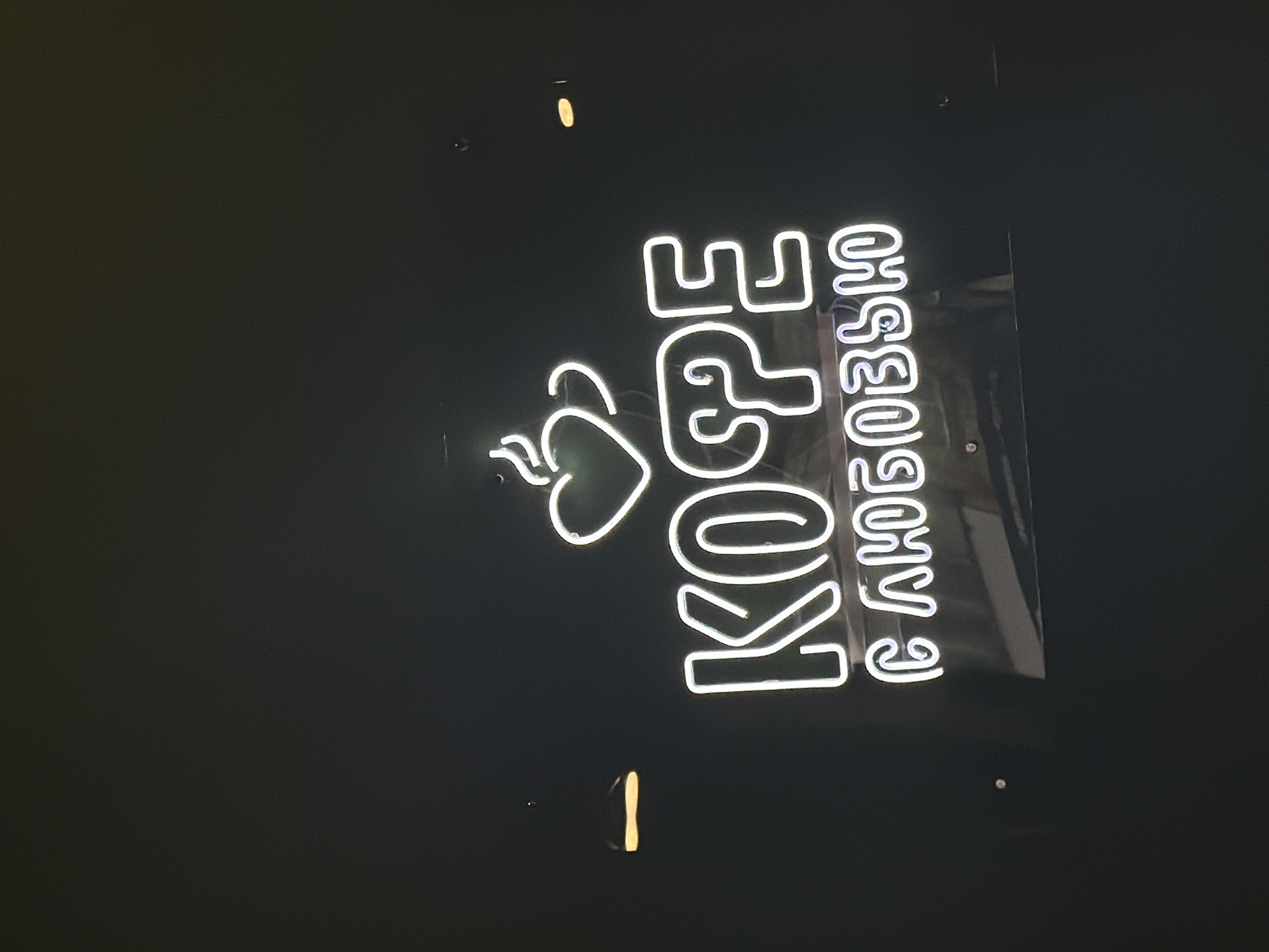 The image shows a neon sign in Russian that translates to "Coffee with Love". The sign includes a stylized image of a coffee bean with a heart shape. The sign is mounted on a dark background, likely a wall. The location is suggested to be in the city of Chaikovsky, Russia, based on the prompt. There are no people visible in the image.