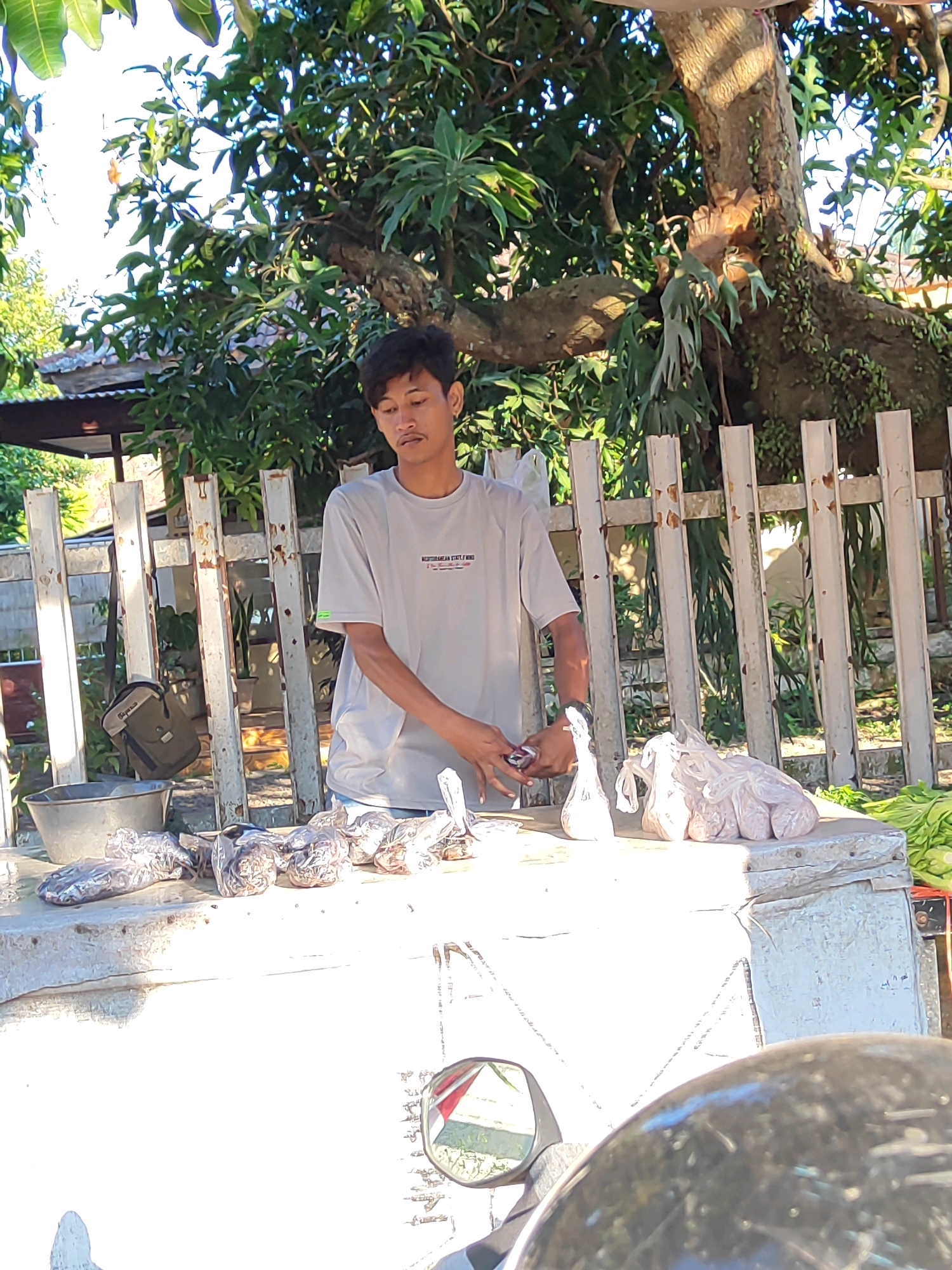 A young man stands behind a table laden with goods, likely for sale, under the shade of a large tree with extensive branches. He is wearing a light-colored t-shirt and appears to be handling some items on the table. The table is covered with a white cloth or surface, and several items are displayed, including what appear to be individually wrapped food products, possibly fish or meat, as well as some leafy green vegetables on the right side of the table. A metal basin is situated on the left side of the table. Behind the table, a white picket fence forms a backdrop. A messenger bag hangs from the fence. The environment suggests an outdoor market or roadside stall. The presence of a motorcycle mirror in the foreground indicates the photo was taken from the perspective of someone on a motorcycle, suggesting a location along a road. The scene likely depicts a vendor in the city of Bondowoso, Indonesia, possibly operating a small business from this outdoor setup.