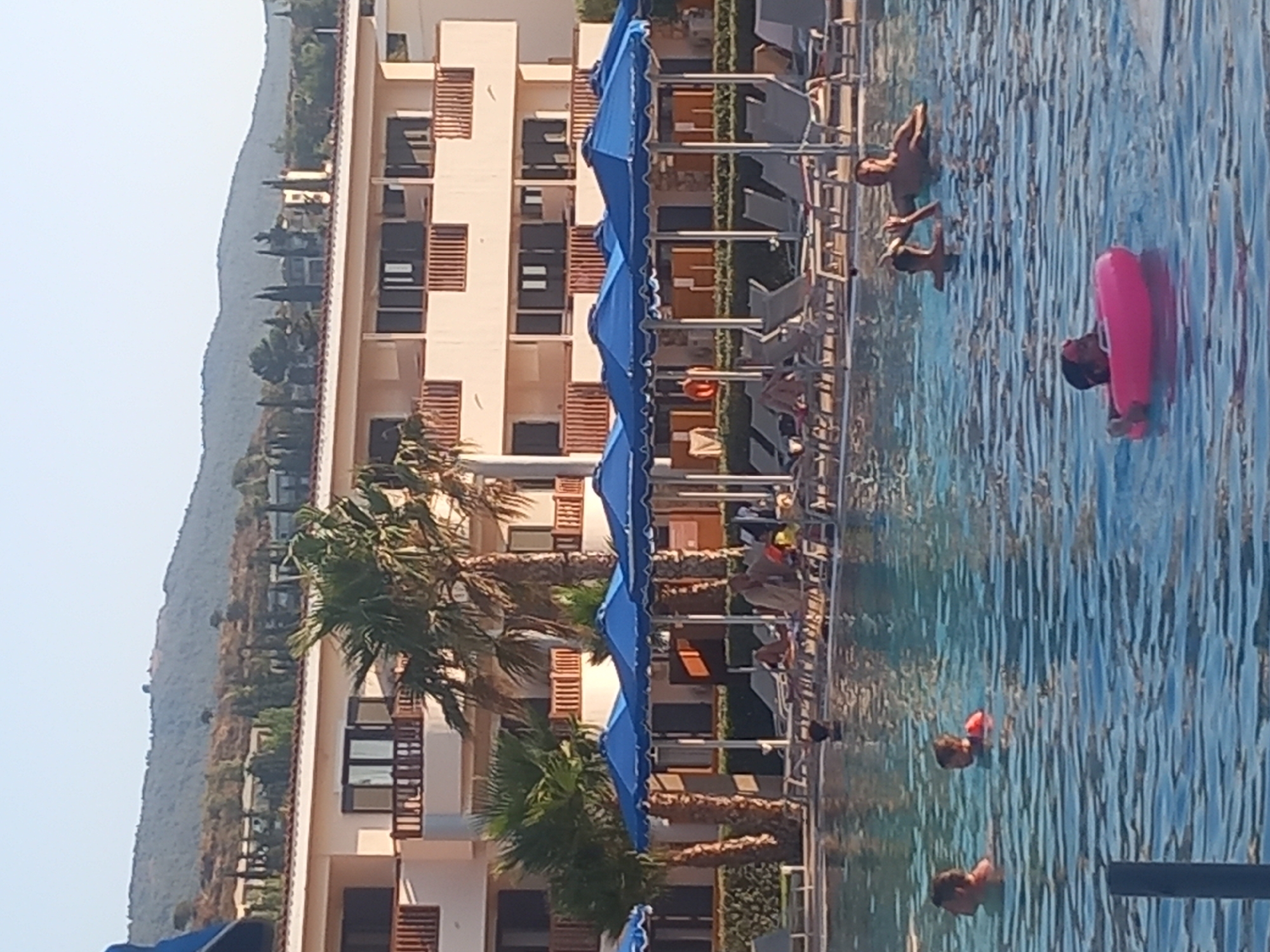 The image depicts a sunny day at a swimming pool, likely at a resort or hotel, with a portion of a multi-story building visible in the background. The building is cream-colored with multiple balconies and dark-framed windows, suggesting Mediterranean architecture. Palm trees are scattered around the property. In the foreground, the blue water of the pool is rippled by movement. Several people are in the pool: a man and a child wearing a red life vest are near the left side, a child in a pink inflatable ring is in the lower right quadrant, and two adults are further back, appearing to interact with each other. Around the pool, numerous lounge chairs are arranged under blue umbrellas, with some individuals relaxing on them. The overall setting appears to be a tourist destination, possibly on the island of Kos, Greece, given the typical resort style and vegetation.