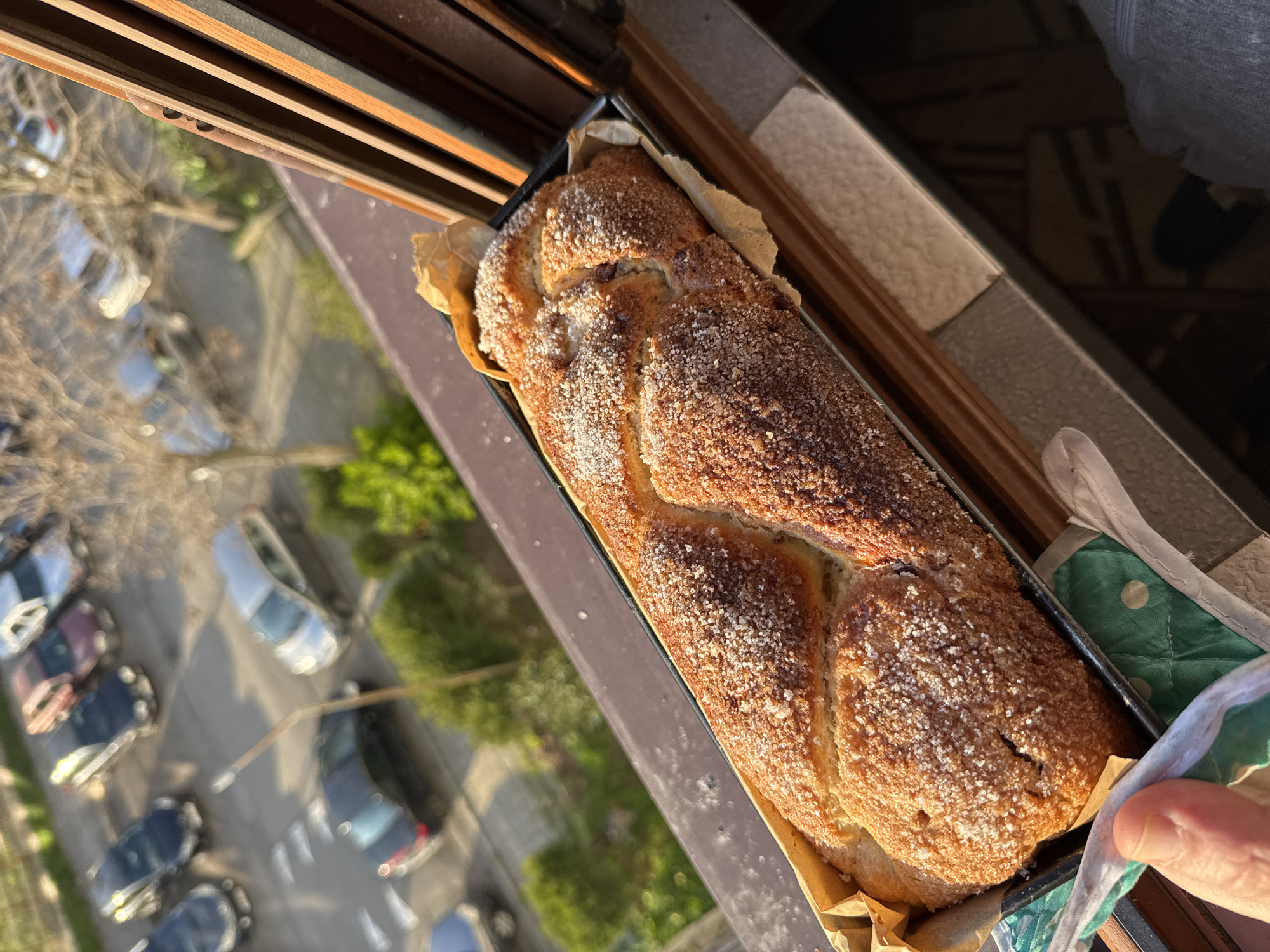 A close-up, top-down shot shows a braided loaf of bread covered in sugar crystals, nestled in a baking pan lined with parchment paper. The pan is placed on a windowsill, with a view of a street scene visible through the window. The street appears to be a residential area, with cars parked along the side of the road and trees lining the street. The location is identified as Burgas, Bulgaria. A hand is holding the pan.