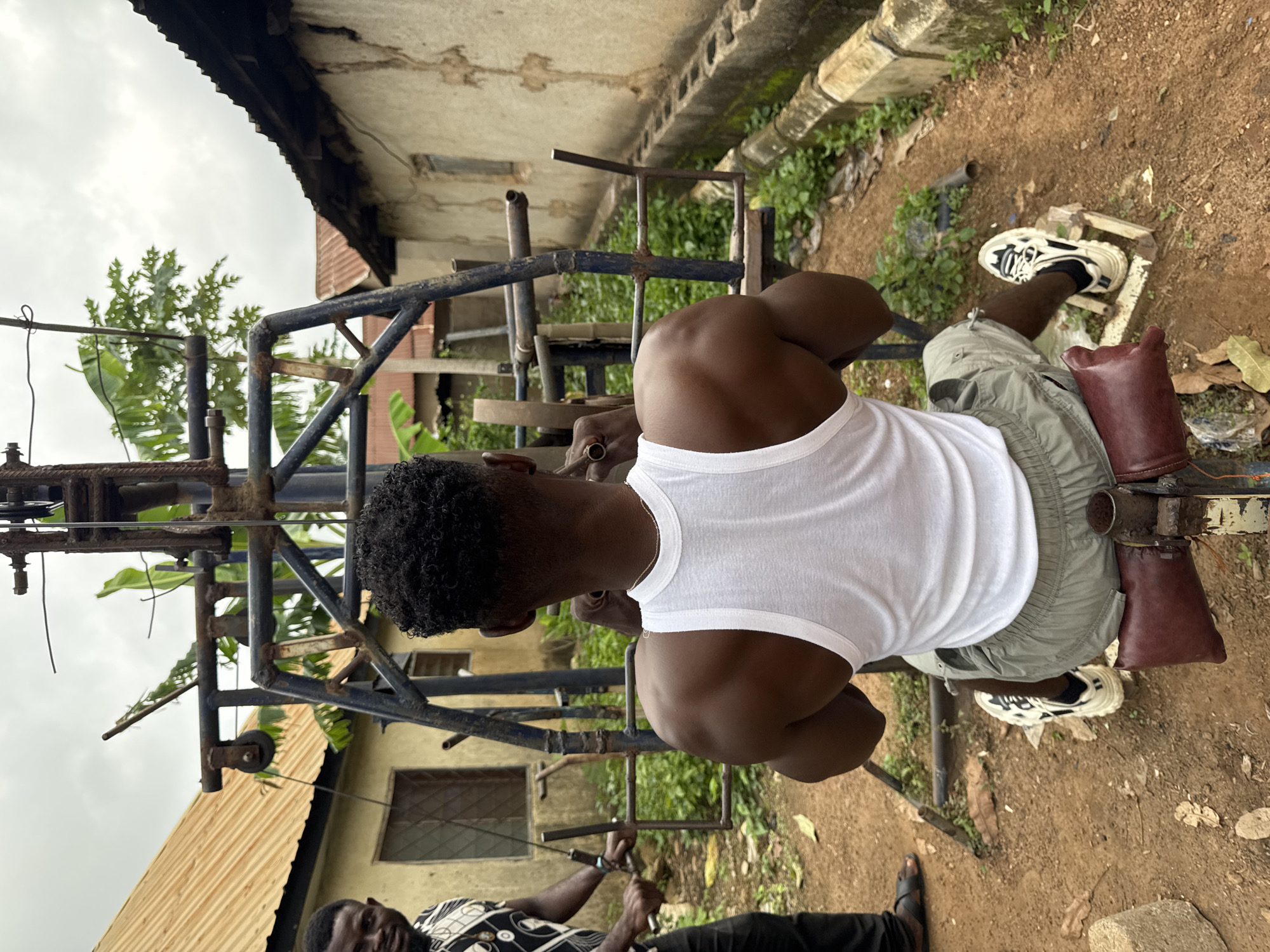 The image shows two men in Akwanga, Nigeria, engaged in a workout. The main subject, facing away from the camera, is a muscular man wearing a white tank top and olive green shorts, actively using a homemade weight machine. He is seated on a red padded seat and his feet are strapped into a wooden platform. His muscular arms are extended and holding the machine's handles.
To the left, another man, partially visible, is also interacting with the weight machine, suggesting he is either assisting or waiting his turn. He is wearing a black and white patterned shirt and dark pants.
The environment appears to be an outdoor space in a residential area. In the background, simple structures with corrugated metal roofs are visible, along with lush green vegetation. The ground is covered with dirt and scattered dry leaves. The scene is well-lit by natural daylight under an overcast sky.