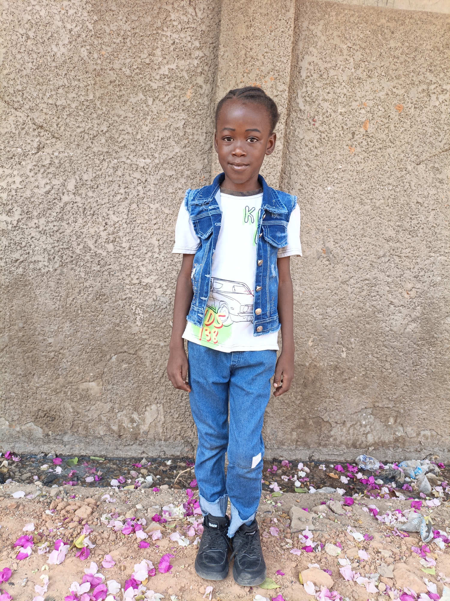 A young Black child stands facing the camera, dressed in jeans, a white t-shirt with a design, and a denim vest. The background is a textured, light brown wall. The ground is covered with pink and white flower petals and some debris. The child is positioned in front of the wall, suggesting an urban or semi-urban environment. Based on the location in Gwagwarwa, Nigeria, this is likely a residential or public space.
