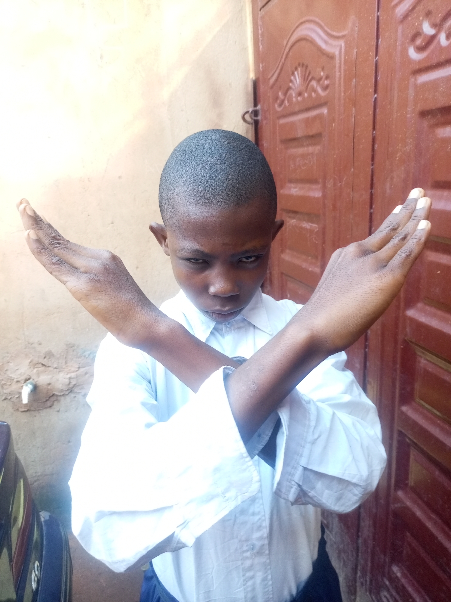 A young boy with dark skin and short, dark hair is standing outside in what appears to be a residential area in Tempure, Nigeria. He is wearing a white collared shirt and dark pants. His arms are crossed in front of his chest, with his hands open and palms facing forward, in a gesture that could signify "stop" or refusal. The background consists of a textured, light-colored wall on the left and a dark red, ornately carved wooden door on the right. A faucet is visible on the wall. The lighting suggests it is daytime.