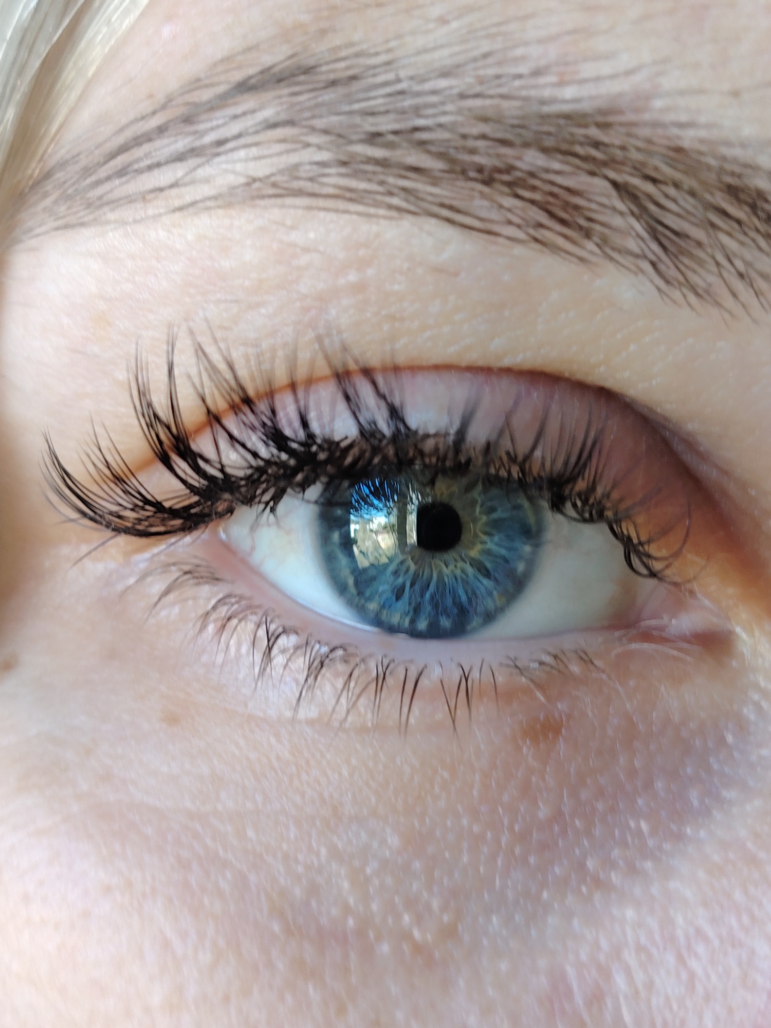 A close-up image shows a blue eye with long, dark eyelashes. The eyebrow above the eye is brown and slightly arched. The surrounding skin is pale and shows fine lines and texture. The reflection in the eye indicates an outdoor environment with trees or buildings. There is no verifiable way to determine the location is Chaikovsky, Russia.