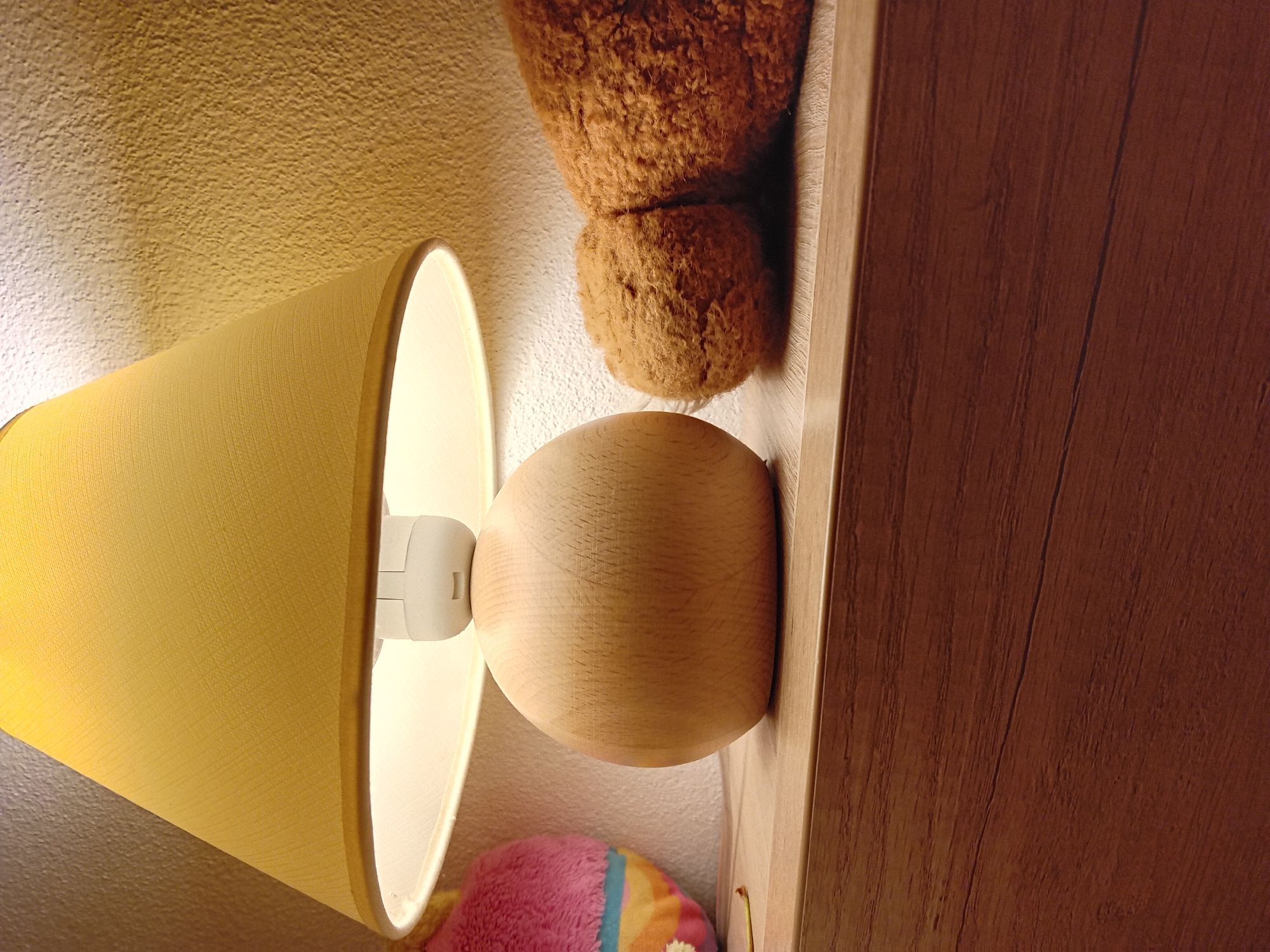 The image shows a close-up of a bedside table with a lamp and two stuffed animals. The lamp has a yellow fabric shade and a wooden base. To the left of the lamp is a pink and rainbow-colored stuffed animal, and to the right is a brown teddy bear. The table surface appears to be made of wood with a light brown finish. The background is a textured wall with a warm, yellowish-brown tone. The lighting suggests a dim, indoor environment. There is no indication of the city of Zuberec, Slovakia, in the image itself.