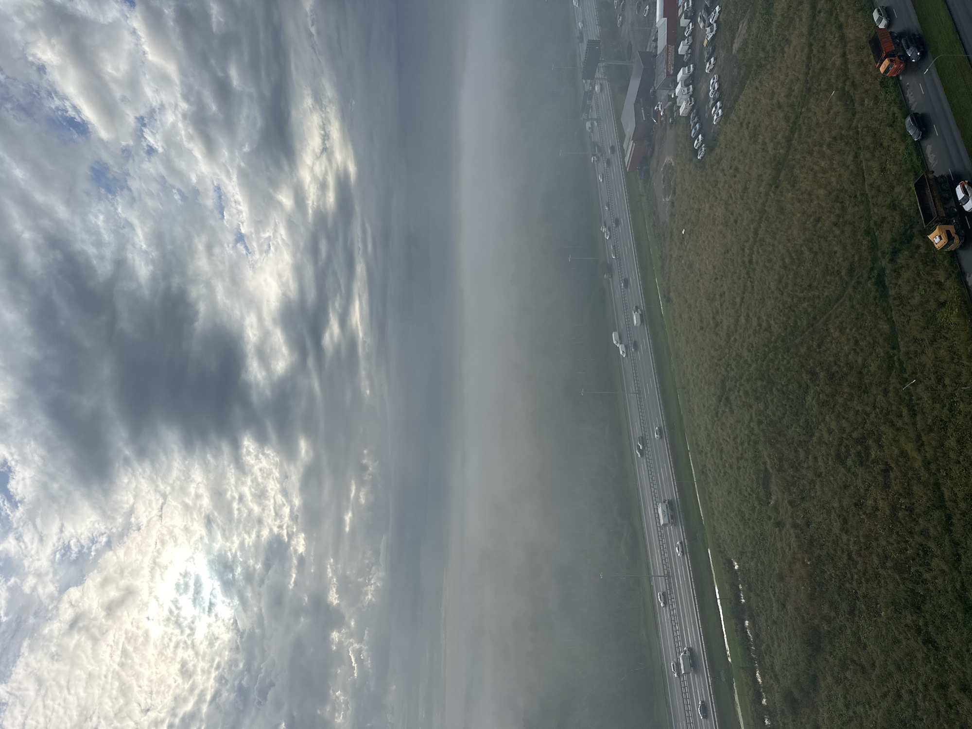This is an aerial, eye-level, medium shot of a foggy highway. The highway is populated with many vehicles. There are also several buildings visible near the highway. The sky is overcast with some bright patches. The overall atmosphere of the image is hazy and somewhat gloomy.