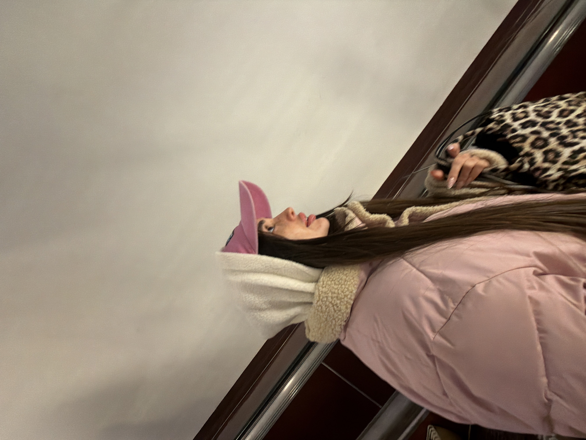 A young woman is riding an escalator in Saint Petersburg, Russia. She is wearing a pink baseball cap, a white hooded jacket with a fur lining, and a pink puffer jacket. She is also carrying a leopard-print bag. The background is a plain white wall and the escalator handrail.