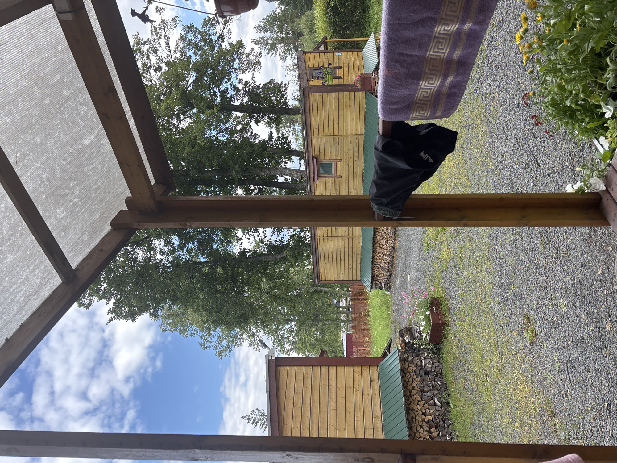 The image depicts an outdoor scene on a cloudy day, viewed from under a wooden and plastic-covered shelter. In the foreground, a wooden railing supports a black garment and a purple towel with a gold geometric pattern. Beyond the shelter, two yellow wooden structures, possibly cabins or small houses, are visible. The structures have green metal roofs. Piles of firewood are neatly stacked against the foundations of these buildings. A gravel driveway or yard surrounds the structures, with patches of green grass and some flowering plants. Tall green trees form a backdrop against the sky, which is partially filled with white clouds. The scene appears to be in a rural or semi-rural setting, consistent with the location in Shuiskaya Chupa, Russia.